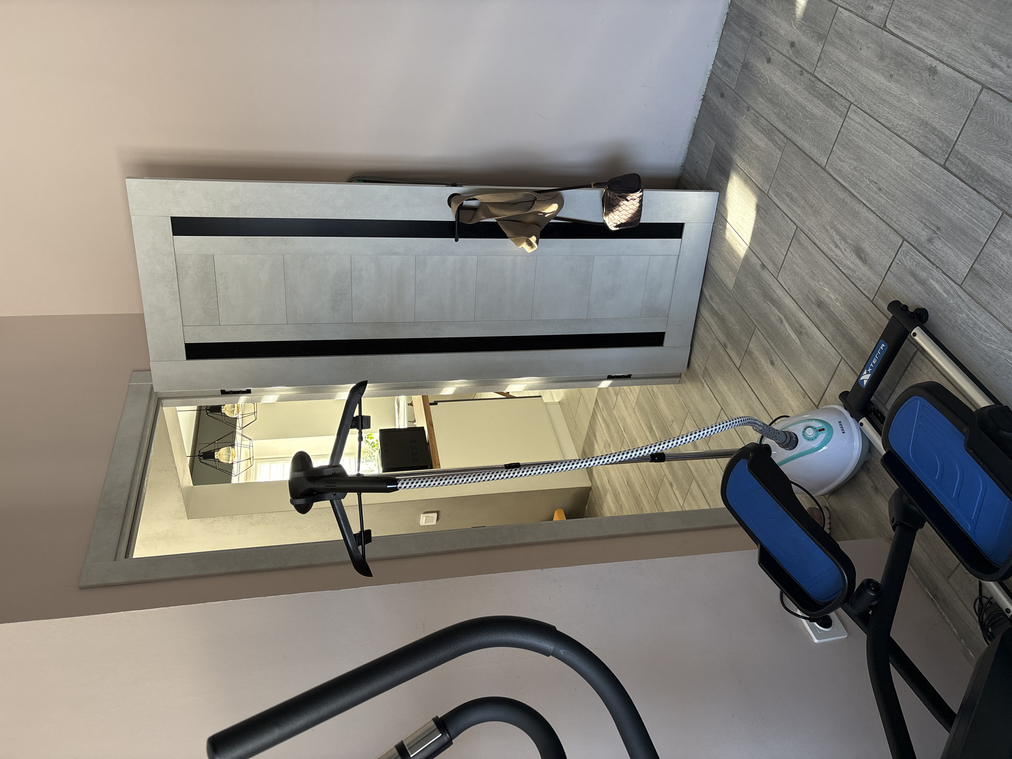 This image depicts an interior residential scene in Votkinsk, Russia, captured from within a room with light pink walls and grey wood-effect tile flooring. In the immediate foreground on the left, a portion of a black exercise machine, identified as an XTERRA brand, with two blue foot pedals, is visible. Adjacent to it, a white electrical outlet is mounted on the wall. Centrally positioned, a KEJI brand floor-standing garment steamer with a white and light blue base, a black telescopic pole, and a black hanger attachment stands on the floor, connected to the outlet. A braided hose with a white and black pattern extends from its base.
To the right, a light grey paneled interior door, featuring two vertical black accent strips, is partially ajar, opening inward to the right. A black horizontal handle is attached to the door, over which a beige garment is draped, with a small, dark brown woven handbag hanging beneath it.
Through the open doorway, an adjoining room, appearing to be a kitchenette or utility area, is visible. This room also has light-colored walls, one of which appears darker grey or green, and features similar grey wood-effect tile flooring. A light wood-toned countertop and white base cabinetry are present, with a dark appliance resting on the counter. Two black industrial-style pendant light fixtures with exposed bulbs hang from the ceiling. A window is discernible in the background, admitting natural light and casting shadows on the floor. No individuals are present in the scene.
