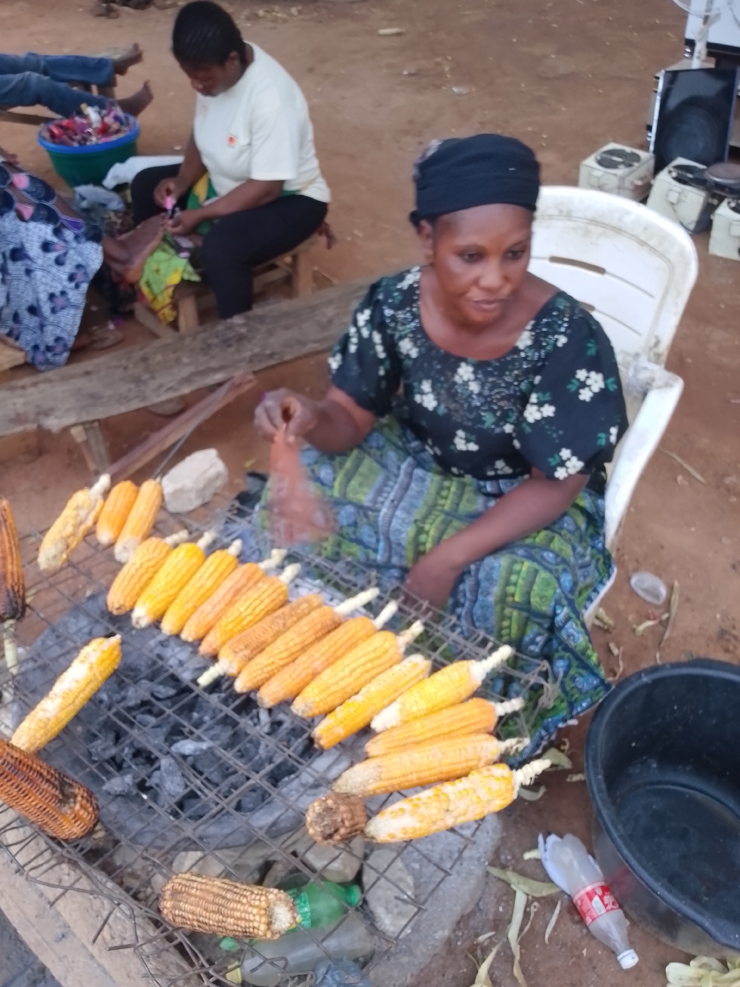 This outdoor scene, set in Sabon Tasha, Nigeria, captures a woman grilling corn over charcoal. The main subject, positioned in the foreground, wears a black headwrap and a dark, floral-patterned blouse with a green and blue patterned skirt. She is seated on a white plastic chair, intently tending to numerous corn cobs arranged on a metal grate above glowing charcoal. Some cobs appear freshly placed and yellow, while others show varying degrees of roasting, turning golden brown or dark. Her right hand is visible, seemingly adjusting or turning a cob.
In the mid-ground, another woman is seated on a low wooden stool, wearing a light-colored t-shirt and black pants. She is focused on an item in her hands, with a blue bucket filled with colorful accessories or craft items beside her. Legs belonging to other individuals are partially visible in the far background, suggesting more people are present.
The ground is unpaved dirt, indicating an informal setting, possibly a street-side vendor stall or a market area. Various objects are scattered around: a large black plastic tub on the right, wooden planks, and another small wooden stool. A green plastic bottle and a red-labeled Coca-Cola plastic bottle are on the ground near the black tub. The "Coca-Cola" logo and text are visible on the bottle. The lighting appears soft, hinting at late afternoon or early evening. The overall atmosphere suggests a communal and busy environment.