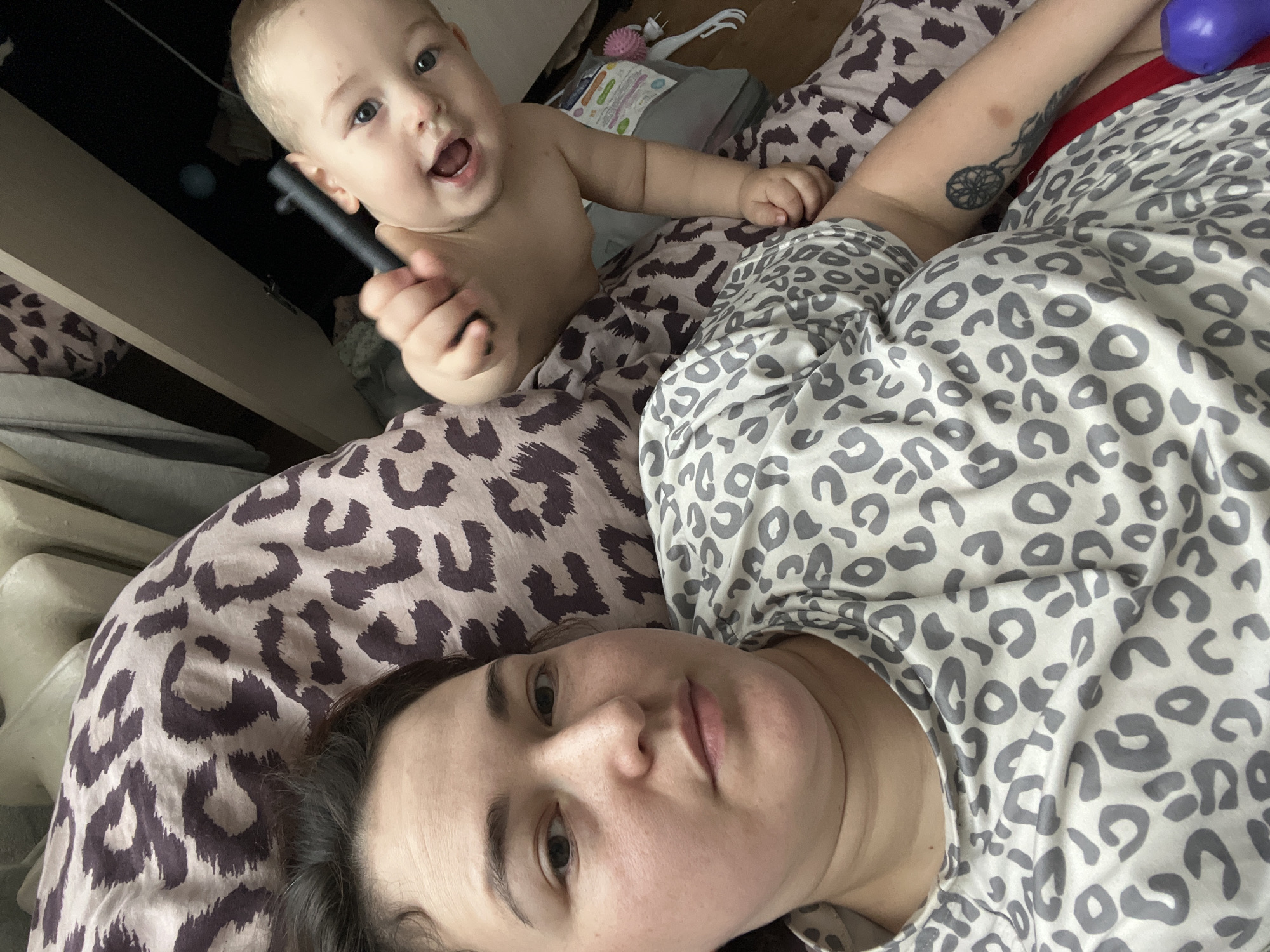 A woman and a toddler are lying on a bed. The woman is in the foreground, her face taking up most of the left side of the frame. She has dark hair and is wearing a grey and white leopard-print t-shirt. The toddler is in the background, on the right side of the frame, reaching towards the camera with a black object in its hand. The toddler has light hair and blue eyes. The bed is covered with leopard-print sheets. There are some clothes and a closet in the background. The scene appears to be indoors, likely in a bedroom. There is no information to determine the location in Chelyabinsk, Russia.