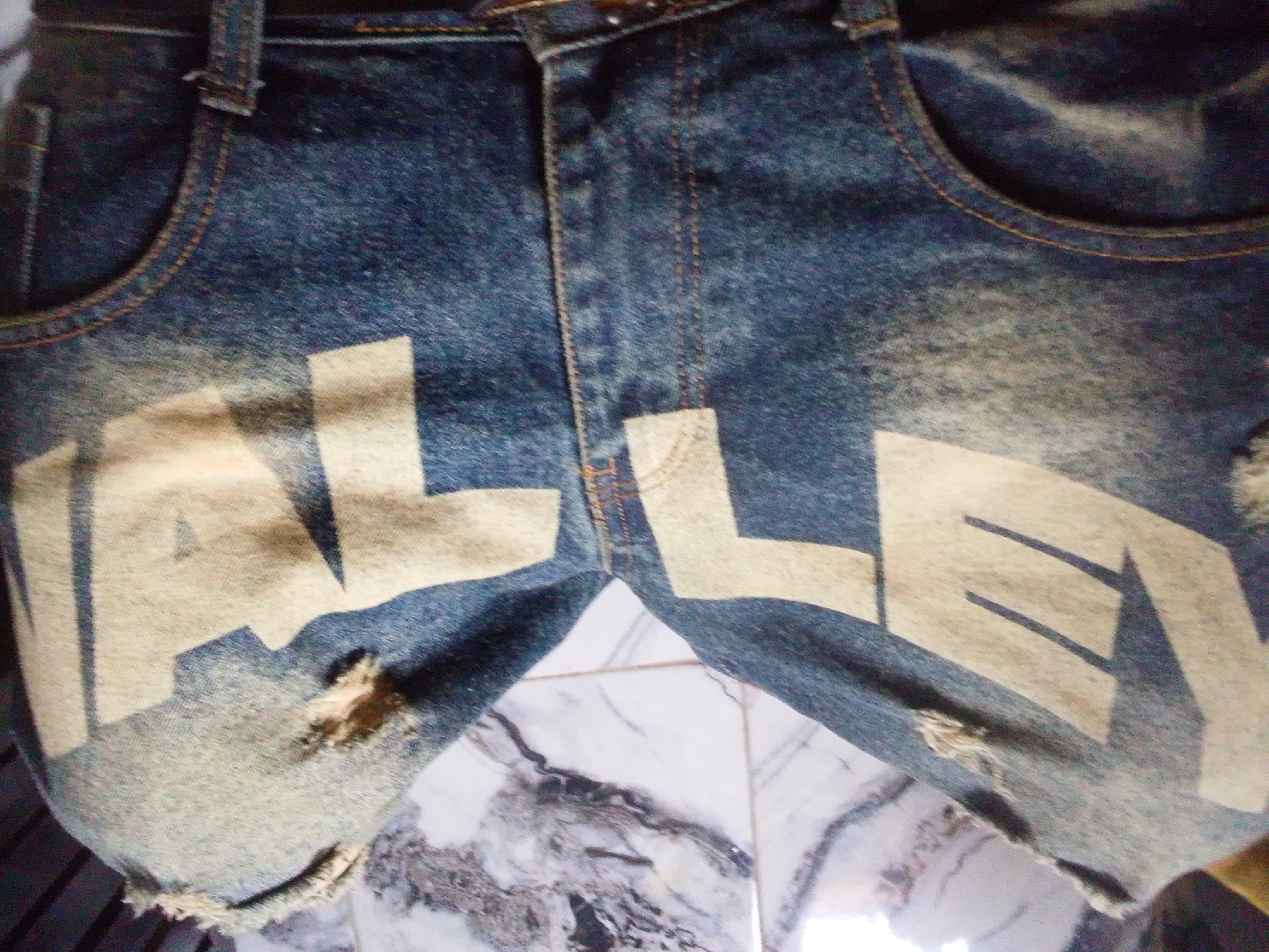 The image is a close-up, low-angle, slightly blurry photograph of the lower front of a pair of distressed denim shorts. The shorts have a faded blue wash with lighter areas and some visible texture. Large, light-colored letters are printed on the fabric. The left leg of the shorts displays the letters "ALLE," with a prominent "A" and "L" visible. The right leg shows the letters "LEY," with the "L" and "E" clearly discernible. There are visible rips and frayed edges on the shorts, particularly near the bottom hem and on the left leg. The shorts appear to be laid flat on a patterned surface, possibly marble or a marble-effect material, with some tiling visible in the background. The lighting is somewhat dim, casting shadows that highlight the texture of the denim.