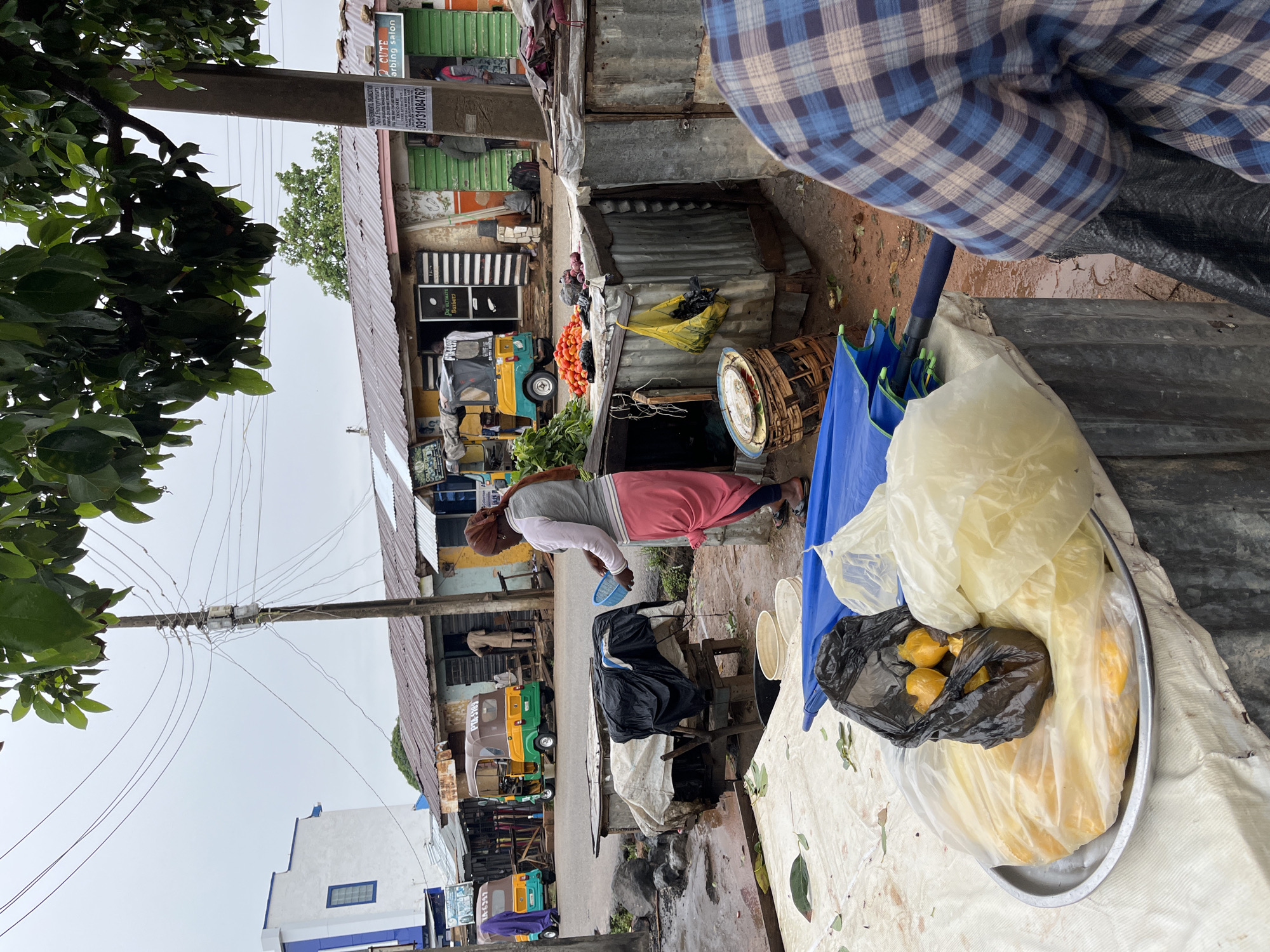 The image depicts a street scene in Jos, Nigeria. A woman in a pink skirt and grey top washes something near a makeshift stall or table laden with plastic bags containing what appears to be food items, including lemons and another yellow produce, all on a silver platter. Next to the platter is a partially visible blue umbrella and woven basket. A person wearing a blue and white plaid shirt is partially visible in the upper right. In the background, several "Keke Napep" auto-rickshaws are parked along the street. The street seems wet or muddy. Above, power lines crisscross near a utility pole with dense green foliage on the left. A sign advertises "2 CUTE arbing salon" and a barbershop alongside phone numbers and the mention of entrepreneurs. The architecture includes structures with corrugated metal roofs, and painted facades such as the building with "PTA-5789" sign.