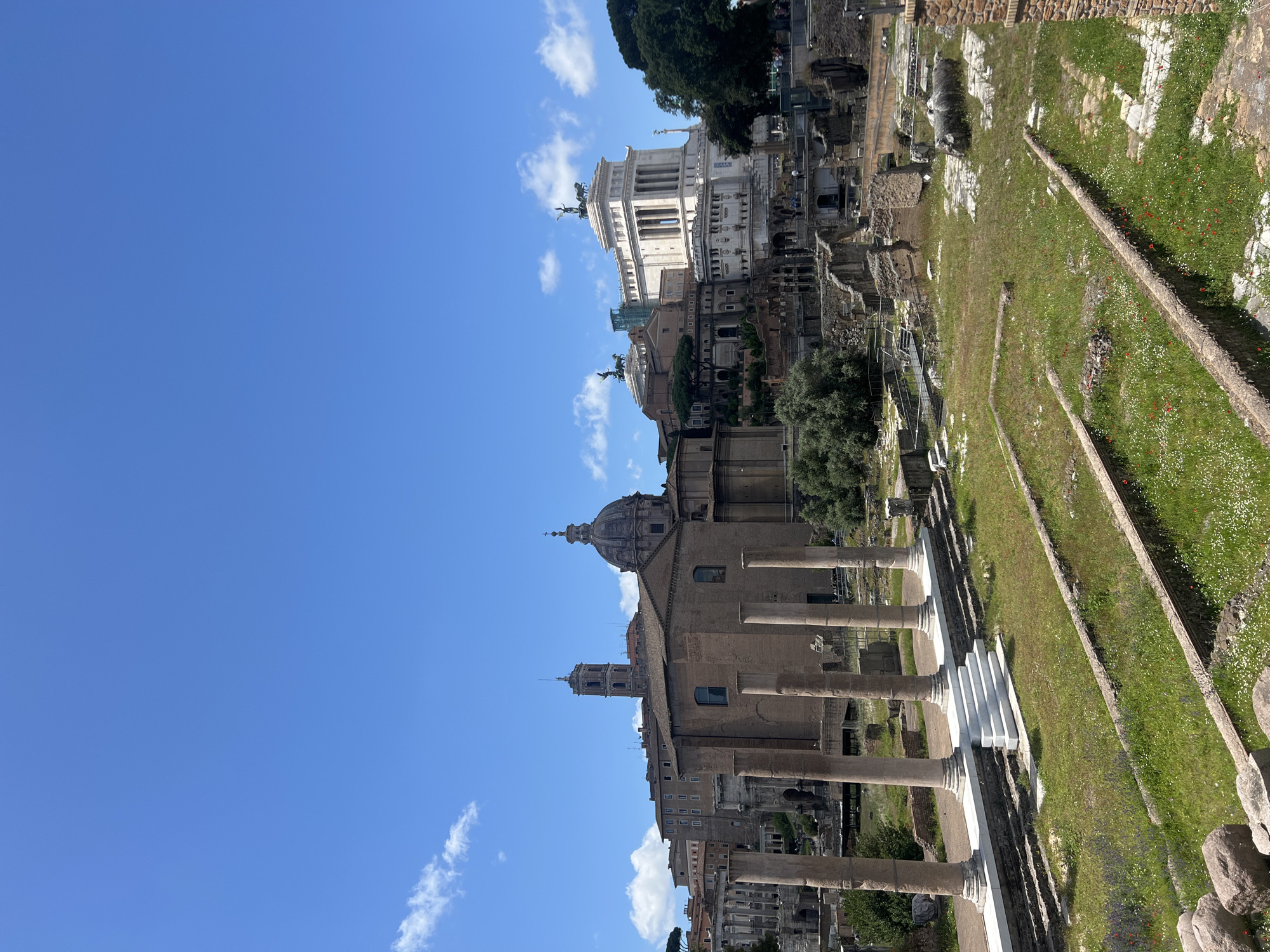 The image shows a view of the Roman Forum in Rome, Italy, under a clear blue sky with scattered clouds. The camera angle is rotated, with the sky on the left and the ground on the right.
In the foreground, ancient ruins and columns stand amidst grassy areas. A path or walkway is visible alongside the ruins, marked with low stone borders.
In the background, several prominent buildings are visible, including a large domed structure and the white marble Victor Emmanuel II Monument.