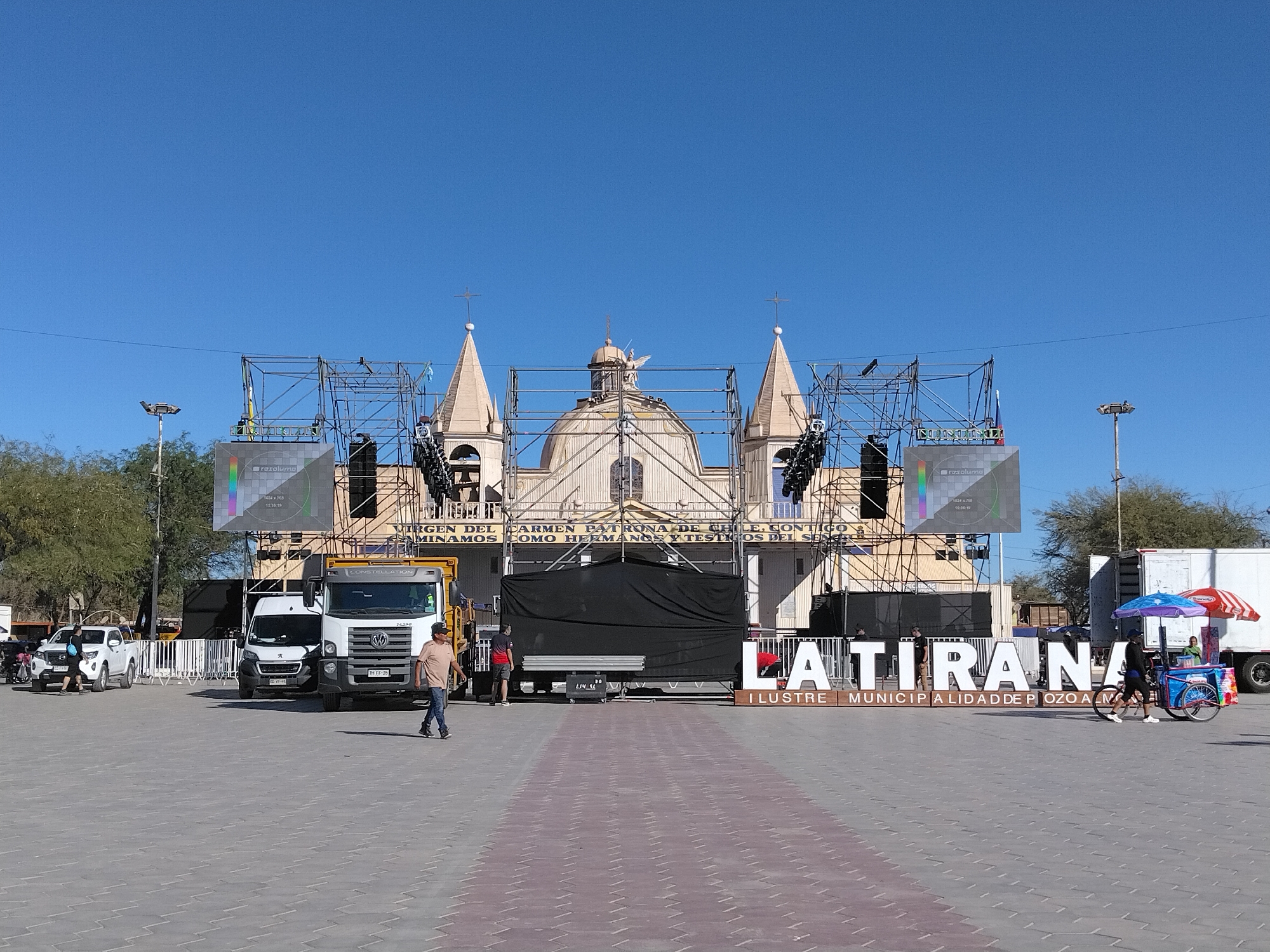 This media file captures an outdoor scene in front of a church, likely in Pozo Almonte, Chile. A large stage with scaffolding and sound equipment is set up in the plaza. Two large LED screens are visible, one displaying a test pattern with the text "resolume" and technical specifications. Banners with Spanish text are also displayed, including "VIRGEN DEL CARMEN PATRONA DE CHILE" and "ILUSTRE MUNICIPALIDAD DE POZO ALMONTE," indicating a religious or civic event.
Several vehicles are present, including a white van and a large yellow truck labeled "CONSTELLATION." A few people are seen in the plaza, with one man walking towards the camera and others working on or around the stage setup. The weather appears clear and sunny, suggesting it's daytime. The overall atmosphere seems to be one of preparation for an event. A colorful cart with umbrellas, possibly selling refreshments, is visible on the right side of the frame. Large, white dimensional letters spelling out "LATIRANA" are a prominent feature of the scene, likely a landmark or part of the event's branding. The ground is paved with interlocking stones.
