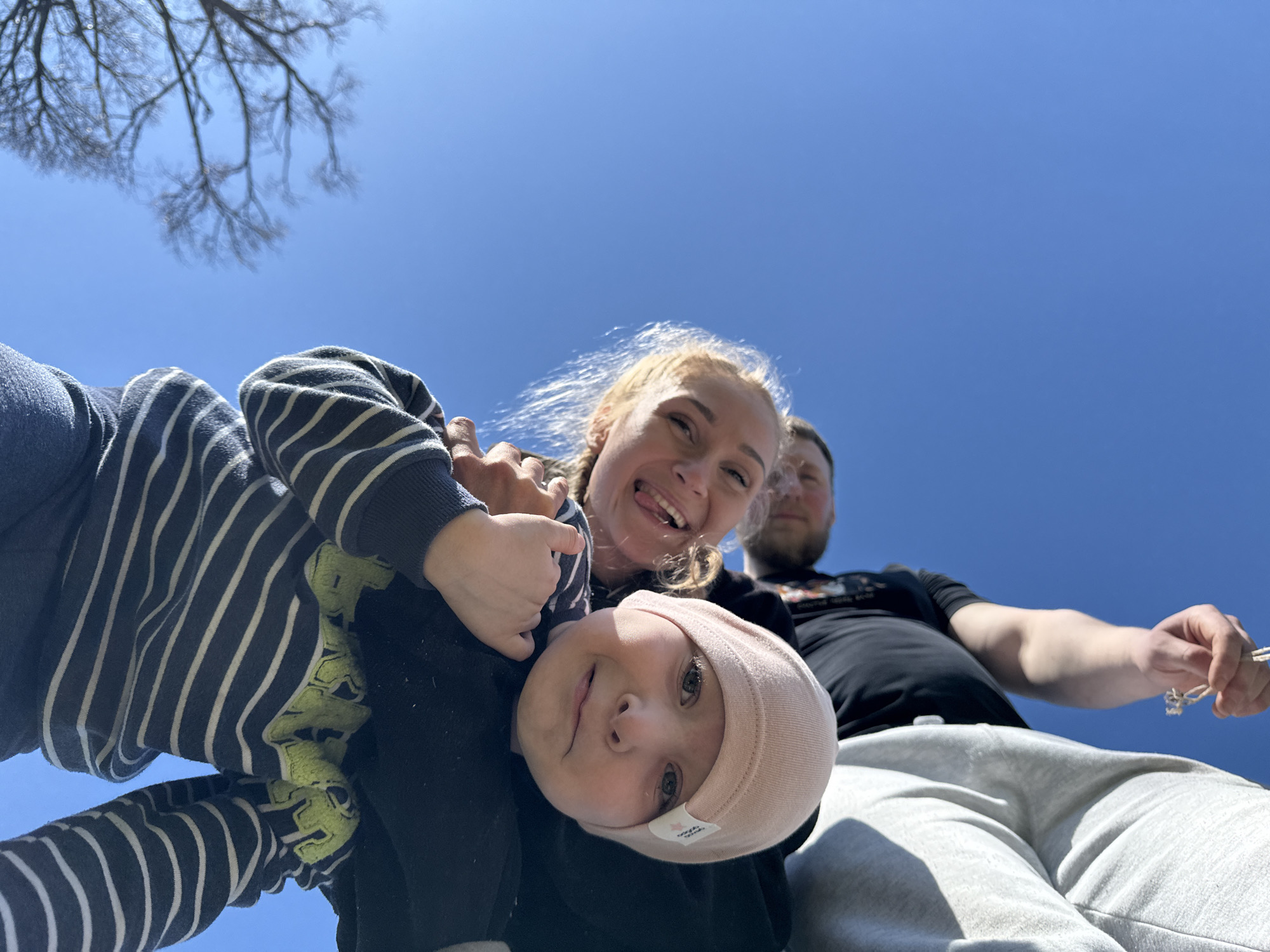 The image presents a low-angle view of three people, likely a family, against a clear blue sky, possibly located in Nizhnyaya Chermoda, Russia given the prompt's location specificity, though there is no explicit evidence of this within the image itself.
A young child, wearing a pink hat, is positioned closest to the camera, appearing to be held aloft. A woman, likely the mother, is behind the child, smiling with her tongue slightly visible. Behind them is a man, likely the father, partially visible and positioned further away.
The child wears a black top over a striped undershirt. The woman wears a black top or jacket. The man wears a black t-shirt and light gray sweatpants, and is holding a thin object between his fingers, possibly a string or small stick.
Above the family, there is a clear, bright blue sky with some bare tree branches visible in the upper left corner. The light suggests it is daytime. The low-angle perspective makes it impossible to determine the surrounding environment or any specific landmarks that would indicate the location within Nizhnyaya Chermoda.