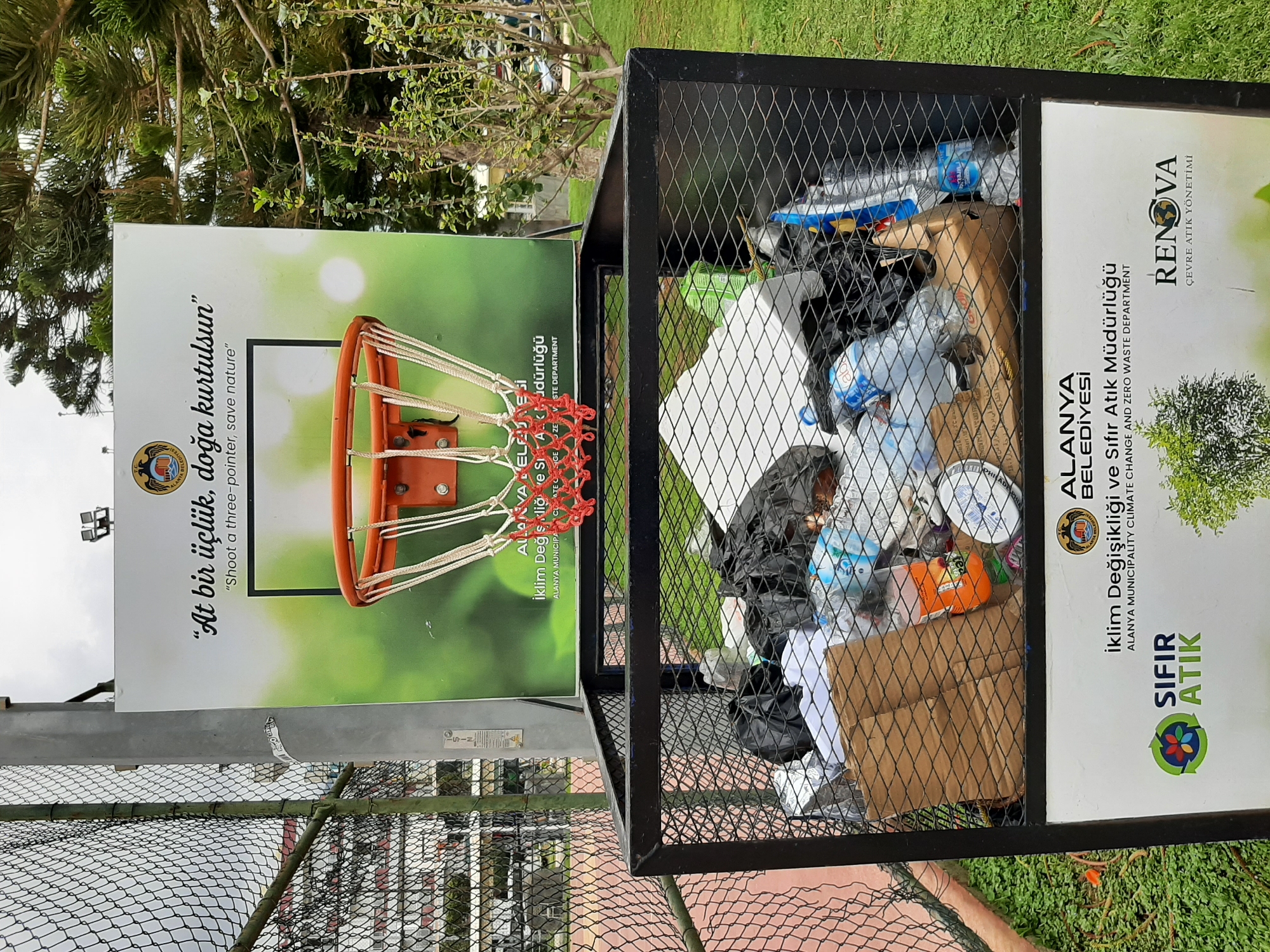 The image depicts a metal trash receptacle in Alanya, Turkey, positioned outdoors next to a poster that features a basketball hoop. The receptacle is full of various types of litter, including plastic bottles, cardboard, and black plastic bags. Adjacent to the trash receptacle is a sign with Turkish text and images of a basketball hoop and a recycling logo. The sign promotes environmental awareness with the slogan "Shoot a three-pointer, save nature." The poster also includes the text "ALANYA BELEDIYESI Iklim Degisikligi ve Sifir Atik Mudurlugu ALANYA MUNICIPALITY CLIMATE CHANGE AND ZERO WASTE DEPARTMENT" and "RENOVA CEVRE ATIK YONETIMI." The background includes grass, trees, and a wire fence, indicating a park or recreational area within the city of Alanya.