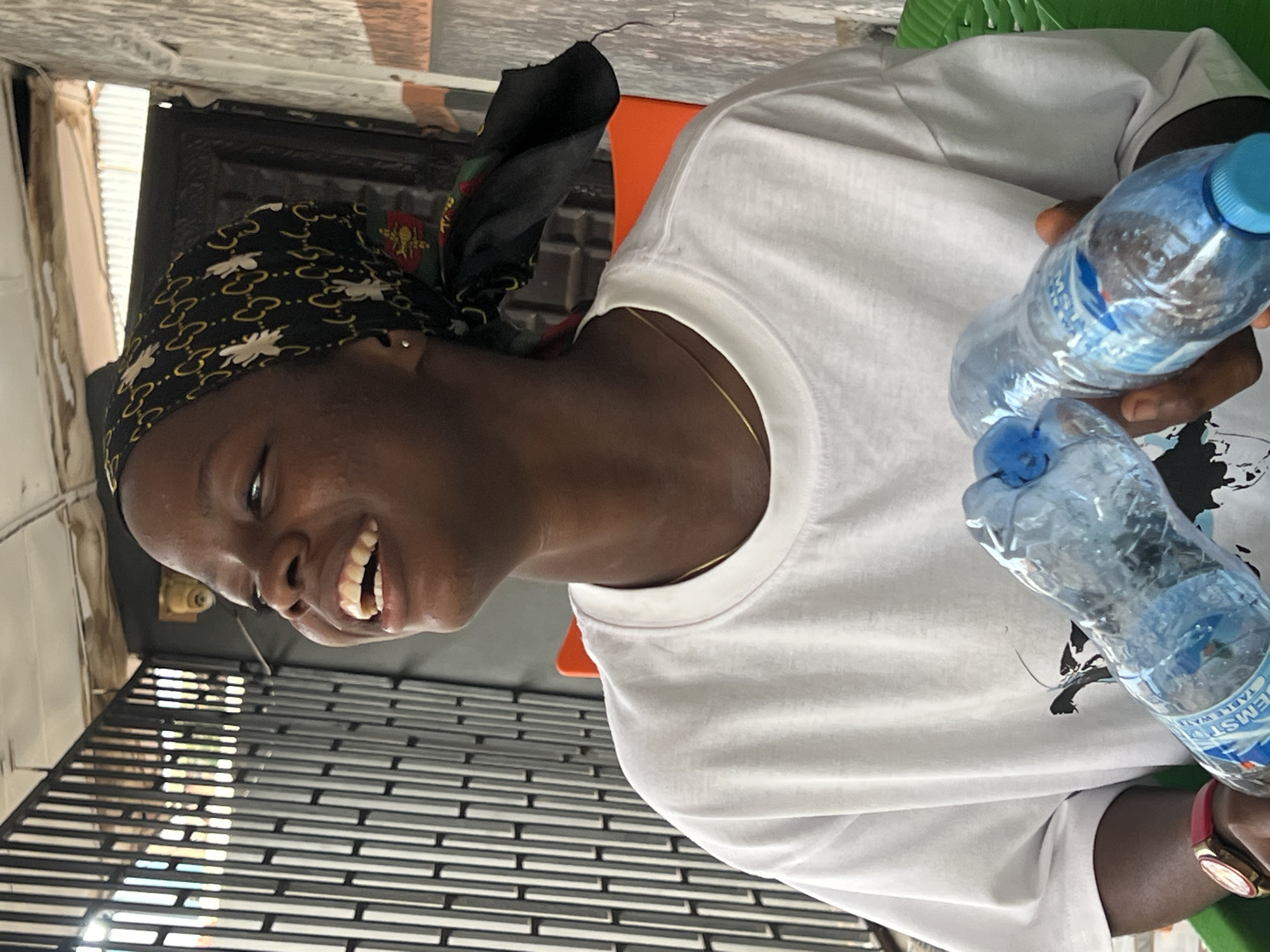 A young Black woman is smiling broadly and holding two plastic water bottles in her hands. She is wearing a white t-shirt and a black headscarf with a gold pattern. She has a gold necklace on. The background shows a metal gate and a wall. The scene is outdoors, possibly in Nkpor, Nigeria, with a corrugated metal roof visible.