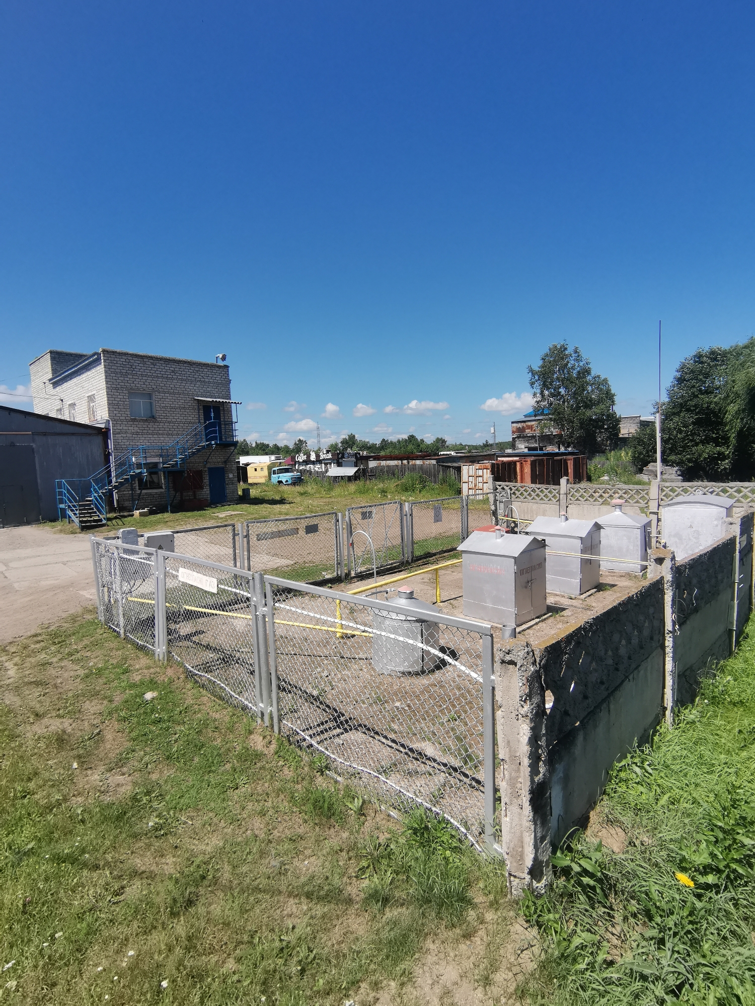 The image depicts an outdoor industrial or utility area under a clear blue sky. In the foreground, a chain-link fence encloses a concrete structure with several metallic, box-like containers, some with peaked roofs. Yellow pipes are visible within the fenced area, along with what appear to be pressure gauges or valves. A weathered concrete wall with visible damage forms the right boundary of the enclosure.
Behind the fence, a light-colored brick building with a flat roof and a blue staircase leading to an upper entrance is situated. Adjacent to this building is a darker, larger structure with a metal roof. Further in the background, various other buildings, some appearing older and constructed from corrugated metal, are scattered across the landscape. A light blue vehicle, possibly a van or small truck, is parked between some of these structures. Trees and greenery are visible in the distance, suggesting a semi-rural or industrial park setting. The ground is a mix of grass, dirt, and gravel.
There are no people visible in the image. The overall scene suggests a functional site, possibly related to gas distribution or a similar industrial process. The location is the city of Vanino, Russia.