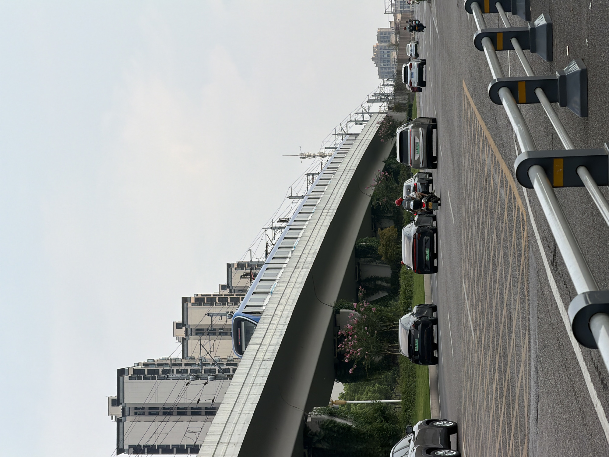 A metro train travels on an elevated track above a city street in Jiangjin, China. The train is blue and white, and its windows are dark. It is traveling to the right. On the street below, several cars and motorcycles are driving. One car is black, and another is silver. A motorcycle with a rider wearing a red helmet is also visible. The street is paved with asphalt and has yellow lines painted on it. In the background, there are tall buildings with multiple stories, some of which are residential. The sky is overcast. The environment appears to be an urban setting.