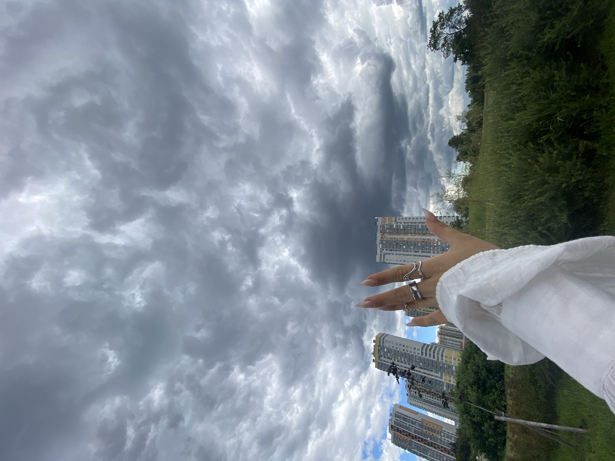 The image captures a dramatic sky filled with dark, heavy clouds over a park-like area with tall, modern buildings in the background. In the foreground, a person's hand, adorned with two silver rings, is raised, with the palm facing the viewer. The hand is covered by a white, loosely draped garment, possibly a sleeve or scarf. The buildings appear to be residential high-rises, consistent with areas of new development in Saint Petersburg, Russia. The greenery in the park suggests an urban green space. The overall mood is one of impending weather, with the contrast between the imposing clouds and the orderly urban landscape.