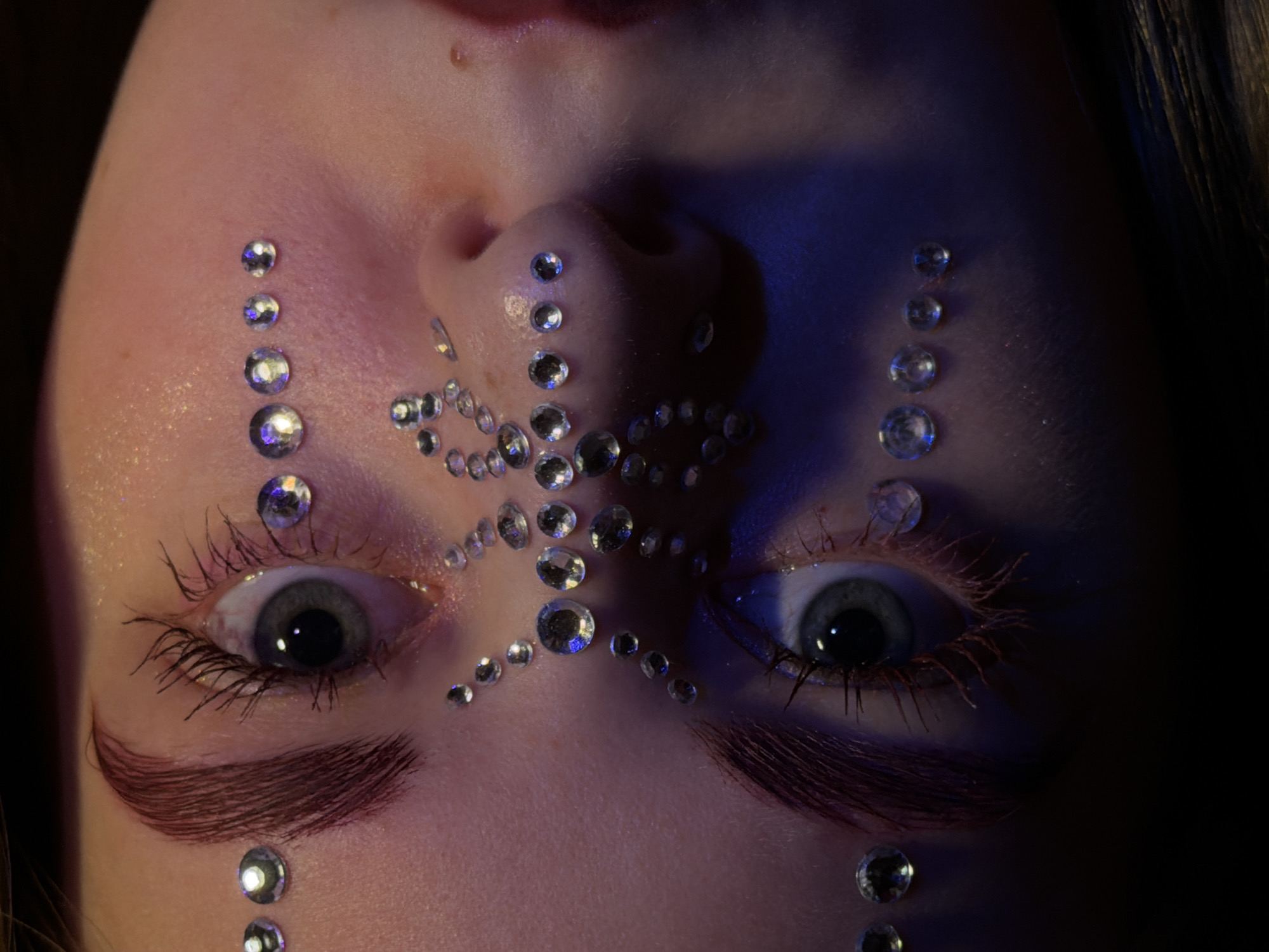 Close-up of a person's face, likely female, tilted upside down, adorned with rhinestones in various patterns around the eyes, nose, and forehead. The eyes are open and appear to be a greyish-blue. The eyebrows are unusually styled and colored brown. The lighting is uneven, with a blue tint on the right side of the face. The image does not depict any location in the city of Perm, Russia, nor actions or interactions.