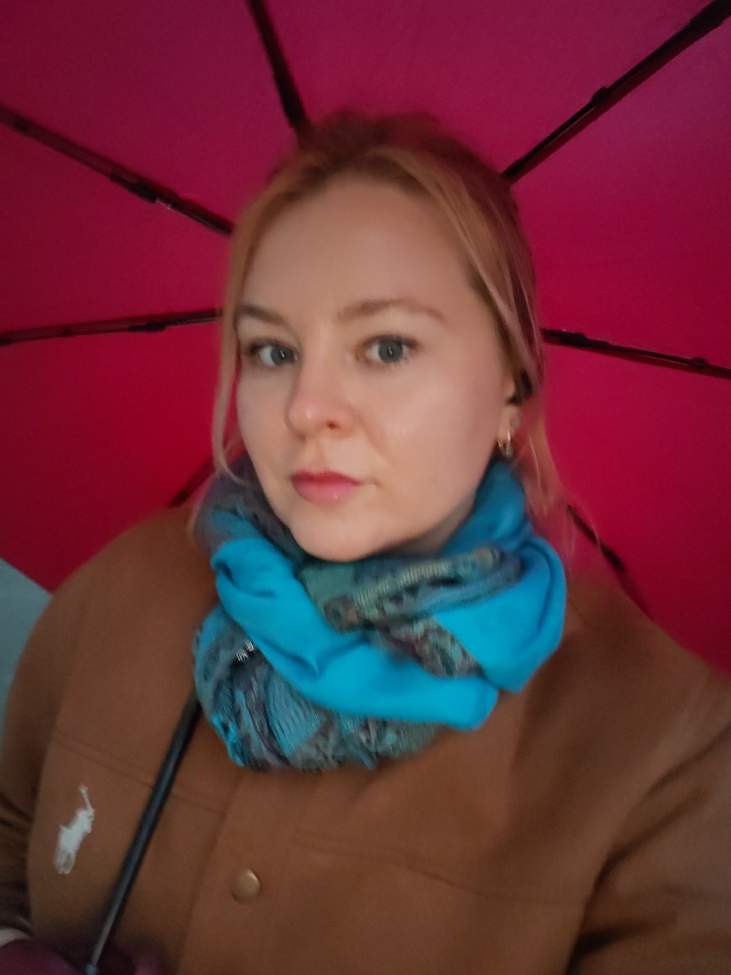 A woman with fair skin and light blonde hair is facing the camera. She has blue eyes and is wearing a bright blue scarf around her neck and a brown coat, which features a Ralph Lauren logo on the left chest. She is standing under a red umbrella. The location is not discernible from the image.