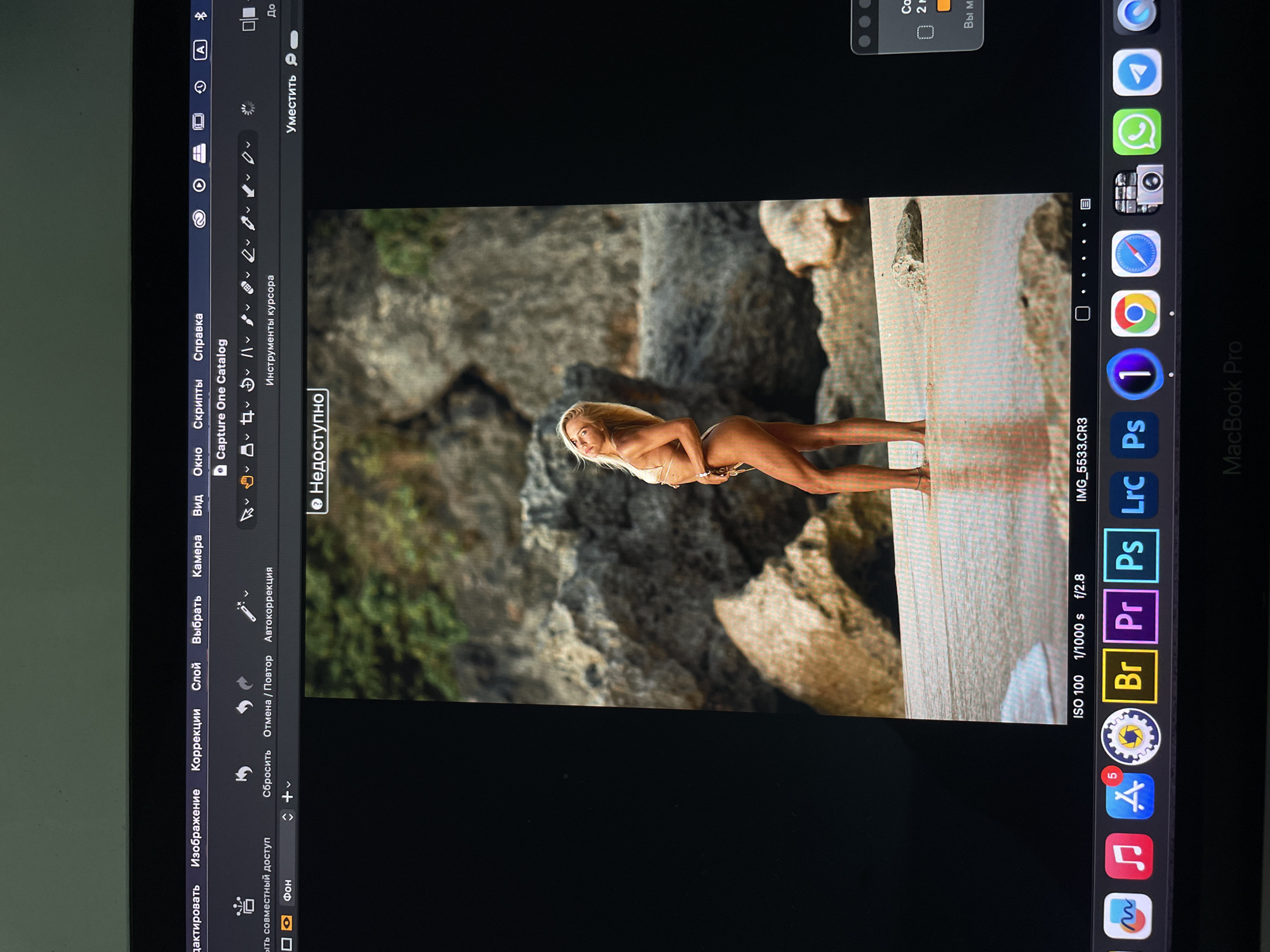 The image is of a MacBook Pro screen. On the screen, there is an open image editing application, "Capture One Catalog," displaying a photograph. The photograph features a woman in a bikini posing on a beach with rocky cliffs in the background. The program shows image settings like ISO 100, 1/1000s shutter speed, and f/2.8 aperture. The MacBook dock at the bottom of the screen displays icons for other applications, including Adobe Bridge, Premiere Pro, Photoshop, Lightroom, and Capture One. Also, there are icons for Safari, Google Chrome, WhatsApp and Appstore.