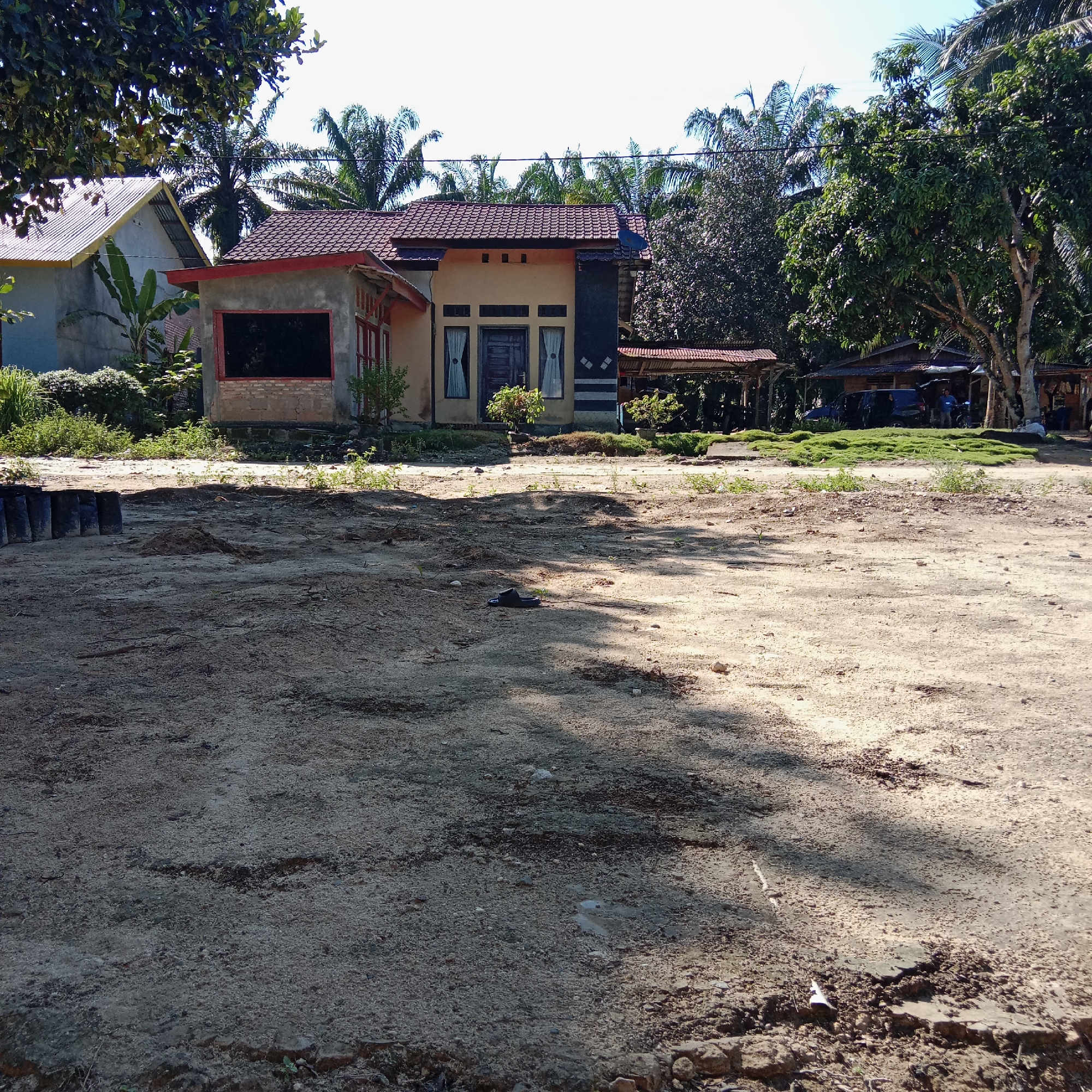 This is a medium-shot, eye-level, outdoor photograph taken in a rural area of Unknown, Indonesia. The scene is dominated by a dirt-covered yard in the foreground, with a house and surrounding vegetation in the mid-ground and background.
The yard is mostly bare earth, with some scattered pebbles and small patches of dry grass. Shadows from trees fall across the ground, indicating it is daytime and sunny. In the lower left of the yard, several black bags are lined up. A single black sandal is placed on the ground in the mid-left foreground.
The house in the mid-ground is a single-story structure with a reddish-brown tiled roof. It has a beige-colored exterior and appears to be constructed with concrete and some brickwork. There are several windows with white curtains and a dark wooden door. To the left of the main house, a smaller, unfinished structure with a reddish frame is visible.
Behind the house, a variety of trees and vegetation can be seen, including palm trees, indicating a tropical climate. In the background, there are some simple covered structures, possibly shelters or workshops, with some vehicles parked nearby. Two individuals are visible standing near one of these structures. The sky is clear and blue.