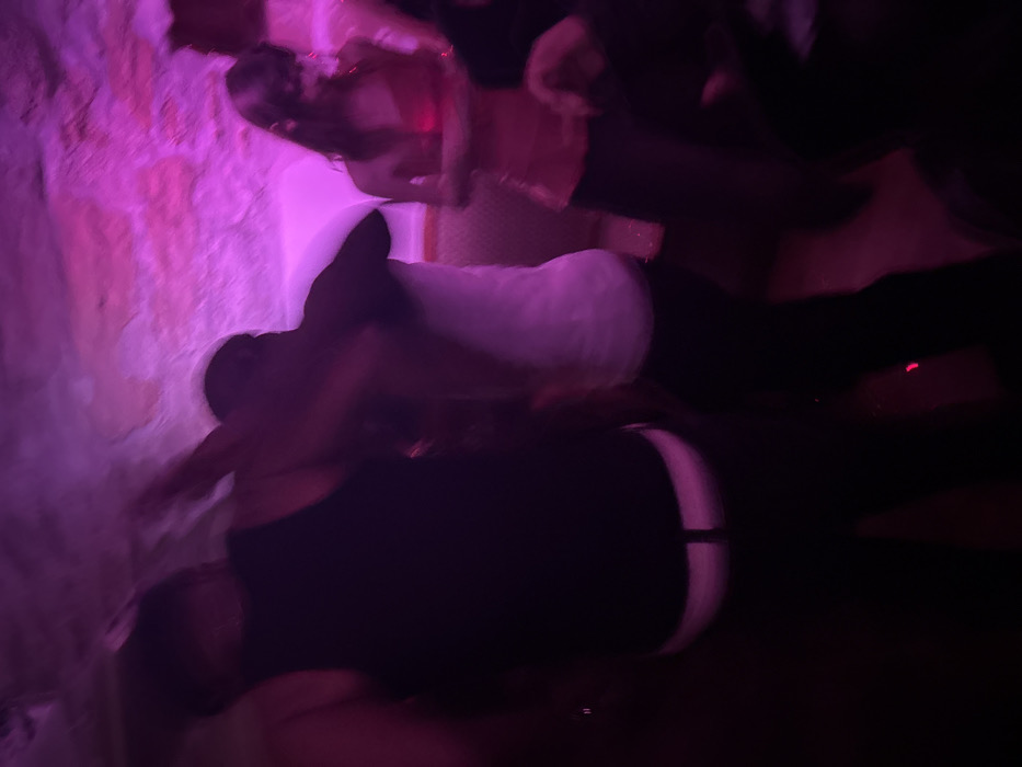 The image displays a blurry, low-light scene captured inside an underground venue in Paris, France, characterized by rough, textured stone walls and ceiling illuminated intensely by pink and purple lighting. Three primary individuals are visible in various states of motion. In the left foreground, a person wearing a black tank top and dark trousers is seen from behind, with a white waistband prominently displayed and their left arm raised. Adjacent, a second individual in a white top and dark trousers is partially visible from behind and to the side, appearing to be in close proximity to the first. To the right, a third person with long hair is depicted wearing a pink or red strapless top, facing slightly away from the viewer with their hands held in front. The overall scene suggests a social gathering or event involving movement or dancing within a cavernous or subterranean space, possibly a repurposed section of the Paris Catacombs or a similar ancient quarry.