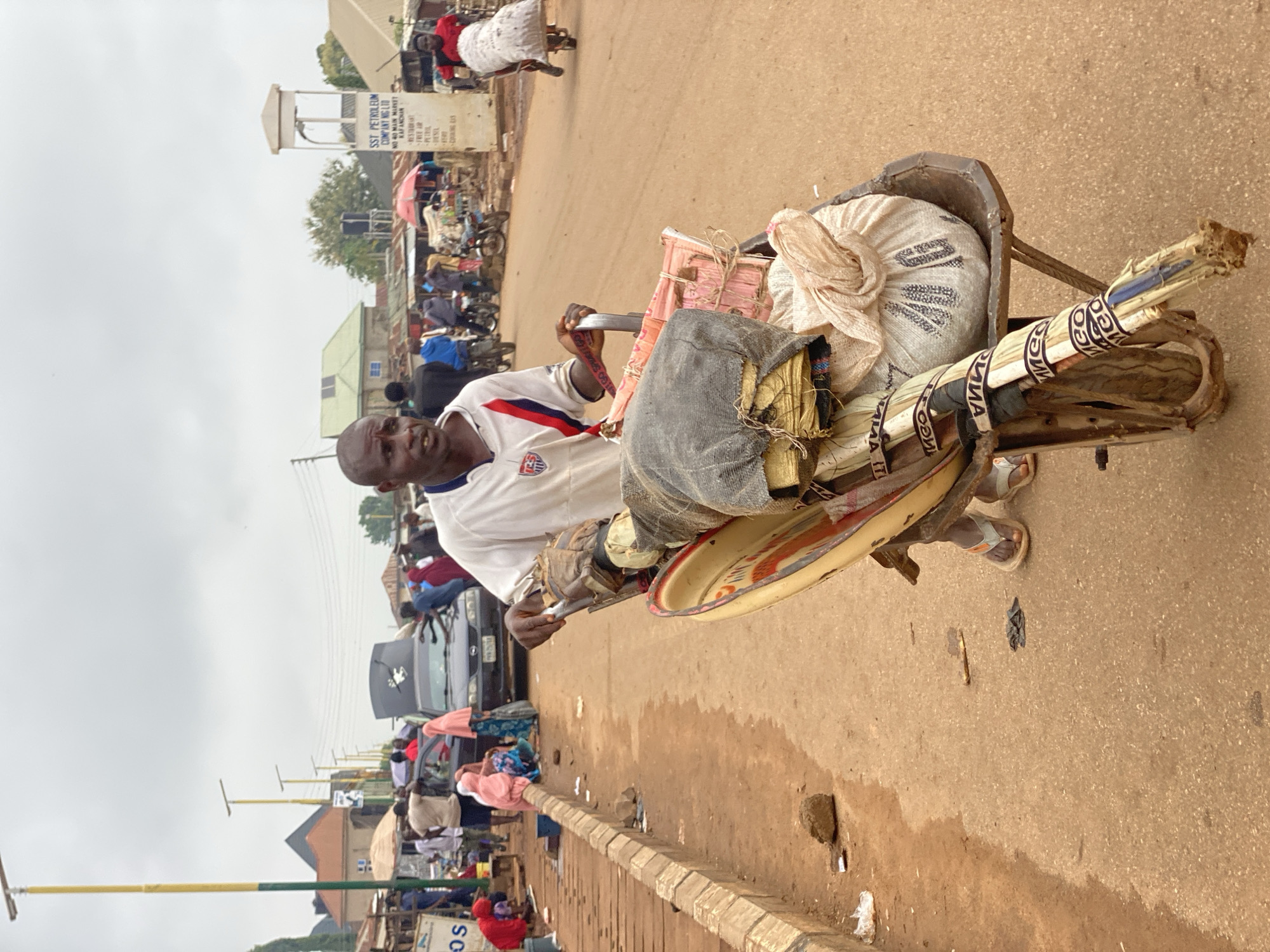 The image depicts a man pushing a wheelbarrow along a street in Kafanchan, Nigeria. The wheelbarrow is loaded with various items, including a large white sack, bundles wrapped in fabric, and a metal tray. The man is wearing a white shirt and sandals. The street scene includes other pedestrians, vehicles, and buildings. A sign for SST Petroleum Company NIG LTD is visible, indicating its location in the city's main market area.