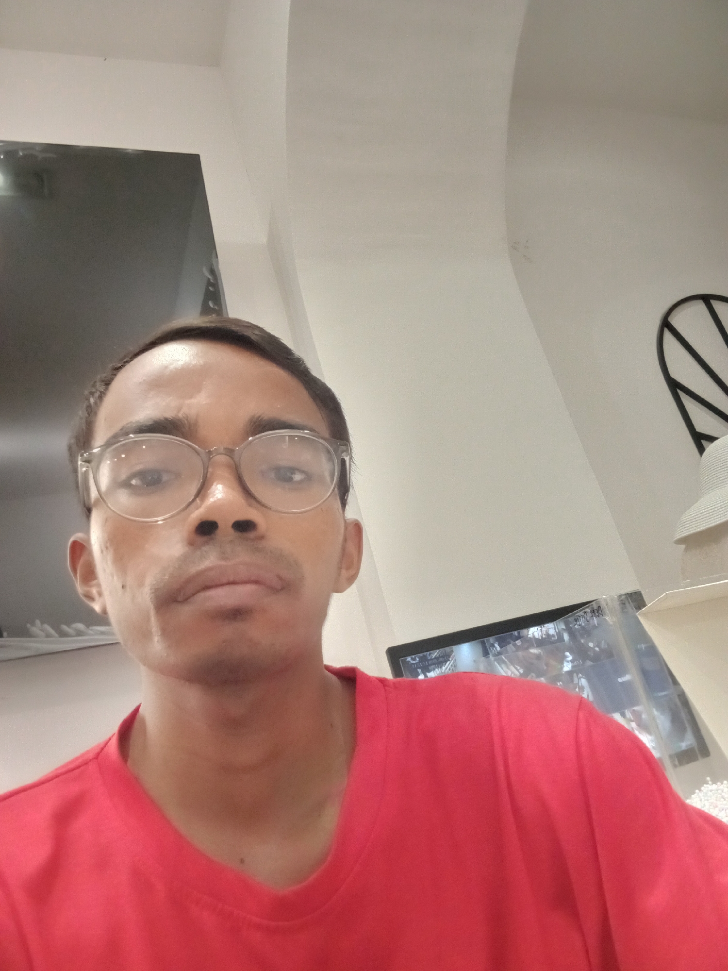 The image shows a medium shot of a young man wearing a red t-shirt and clear-rimmed glasses. He has short, dark hair and a faint mustache. He is looking directly at the camera with a neutral expression. The background is out of focus and appears to be an indoor setting with a white wall and a ceiling that has an arched design. To the left, a dark rectangular object, possibly a large screen or mirror, is visible. To the right, a monitor displaying an image can be seen, along with what appears to be a white hat. The overall lighting suggests an indoor, possibly office or home environment. The location is specified as the city of Larangan, Indonesia.