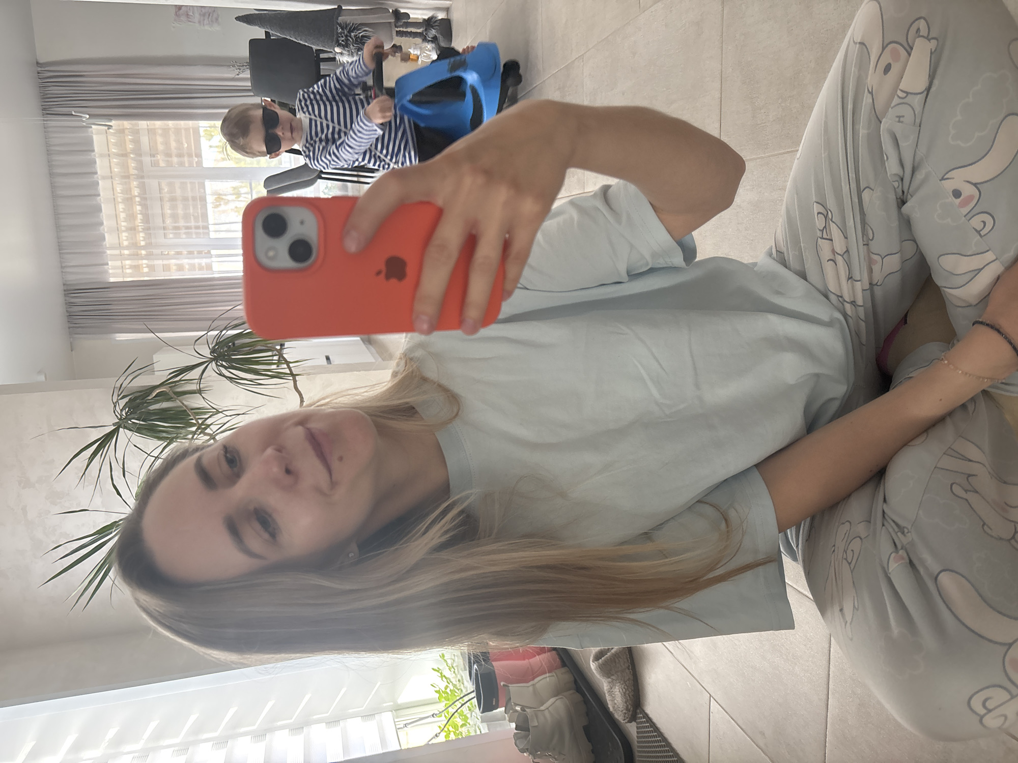 A woman takes a selfie indoors, likely within a home in Chaikovsky, Russia. She is seated on a light-colored tiled floor, wearing light blue pajamas patterned with cartoon bunnies and a light blue t-shirt. Her long blonde hair is visible around her face as she holds a red iPhone, clearly showing the Apple logo, to capture the image. She has a subtle smile.
In the background, a child wearing dark sunglasses and a blue and white striped long-sleeved shirt is seated in a chair, with a bright blue object, possibly a booster seat or toy, positioned in front of them. The room is illuminated by natural light from a large window fitted with light-colored curtains or blinds. A tall potted plant stands near the woman's head. A small letter 'H' is visible on one of the bunny patterns on her pajamas. The image conveys a casual, daytime domestic scene.