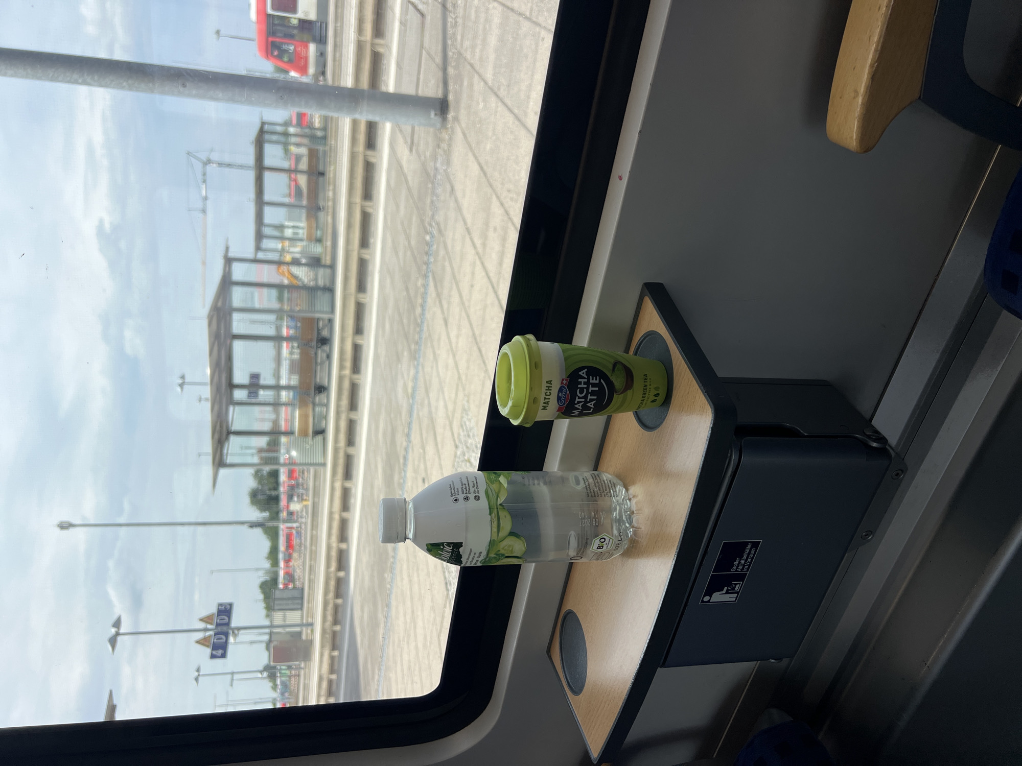 The image, taken from inside a train at the Mühldorf a. Inn, Germany train station, shows a tray table with a bottle of water and a matcha latte cup. Outside the window, train tracks and platforms are visible, along with a red and white train. The sky is cloudy, and there is a crane in the distance. The tray table has a built-in trash receptacle with a German symbol and text indicating it is a large trash bin in the anteroom.