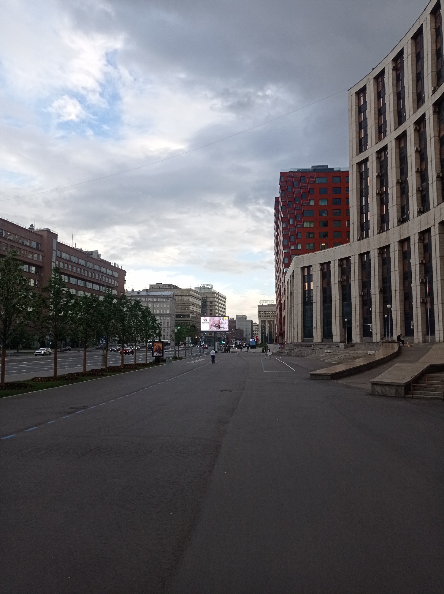 The image captures a wide-angle view of a city street with modern architecture under a cloudy sky. The scene depicts a street in Moscow, Russia, likely during the daytime, with overcast conditions contributing to a muted light.
The foreground is dominated by an asphalt road, with a blue line marking a lane. To the left, a row of young trees with sparse foliage lines the sidewalk, which is separated from the road by a curb. Beyond the trees, a multi-story brick building with numerous windows stretches along the street. Further down the street, cars are visible on the road, suggesting ongoing traffic.
On the right side of the image, a grand building with a facade of vertical white panels and dark windows stands prominently. This building features an elevated entrance with concrete steps and railings. To its left, a striking, modern red building with a distinctive geometric pattern of windows rises high. Between these structures and further down the street, other buildings of varying architectural styles and heights are visible.
In the distance, a large electronic billboard displays an advertisement with some Cyrillic text, although it is not entirely legible. Several figures are scattered along the street and sidewalk. Some individuals are walking, and a few appear to be sitting on the steps of the building on the right, possibly taking a break. The presence of people, cars, and buildings indicates an urban environment with daily activities underway. The overall mood is subdued, possibly due to the cloudy weather and the imposing nature of the urban landscape.