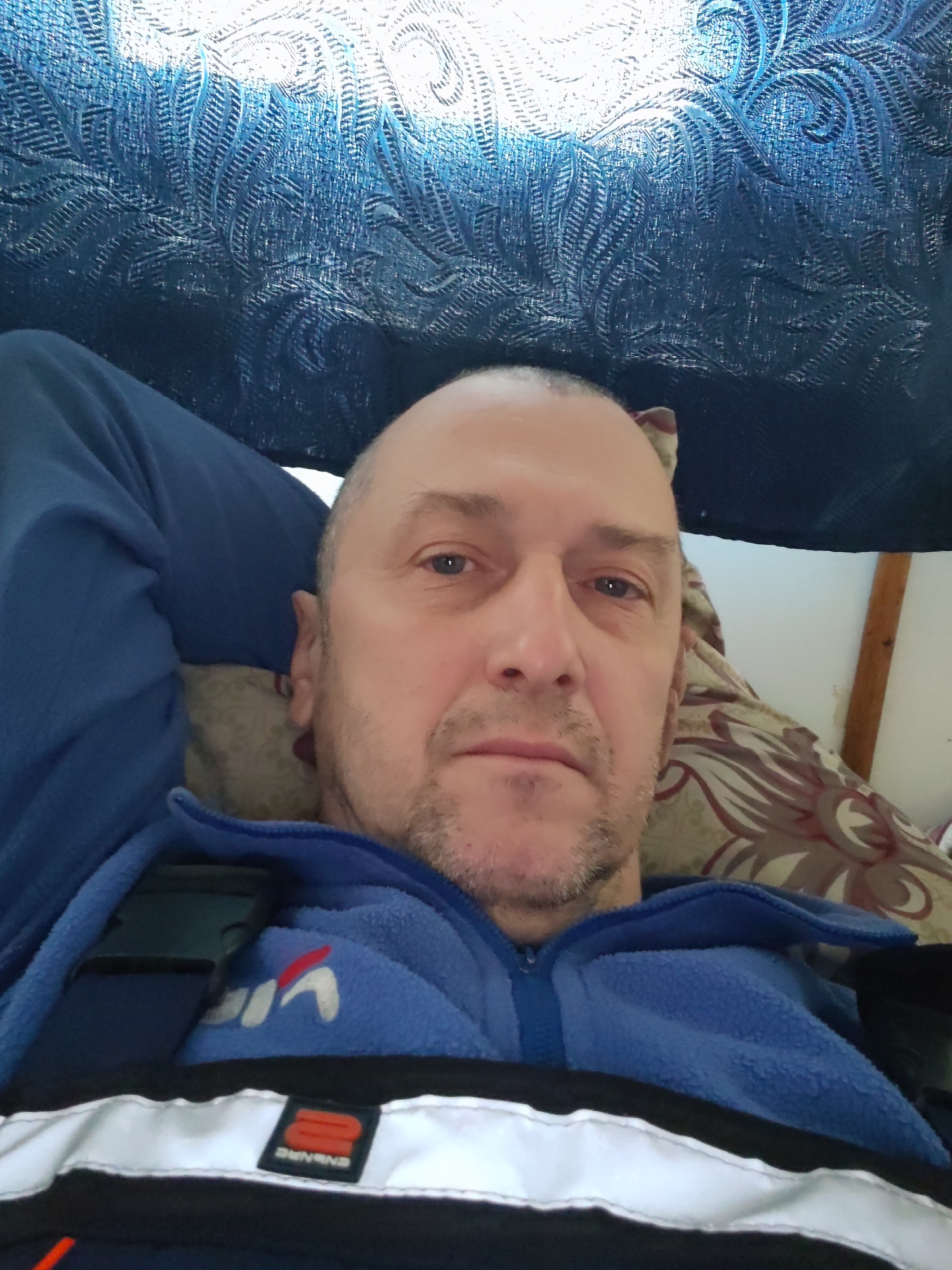 A middle-aged man is captured in a close-up selfie, lying down and looking directly at the camera with a neutral expression. He has a bald head with short stubble on his face and light-colored eyes.
He is wearing a blue fleece jacket, partially zipped, over a dark blue garment that displays a white and red stylized logo resembling "VIA". Across his chest, a black strap with a buckle is visible, suggesting it might be part of a harness or a bag strap. Below this, a black band with a white reflective stripe runs horizontally. On this stripe, a small, rectangular black and red/orange label is attached, featuring the word "ENSHINE" printed upside down in white.
The man's head rests on a pillow with a patterned cover, displaying brown and red swirling or floral designs. Behind and above him, a heavily textured, dark blue fabric with a wavy or leafy embossed pattern creates the background, possibly a blanket or part of an interior covering. To the upper right of his head, a light-colored wall and a wooden beam or frame are visible. The lighting is bright, particularly highlighting the textured blue fabric above him. The overall scene suggests a moment of rest, possibly indoors, with the man's attire hinting at workwear or practical gear.