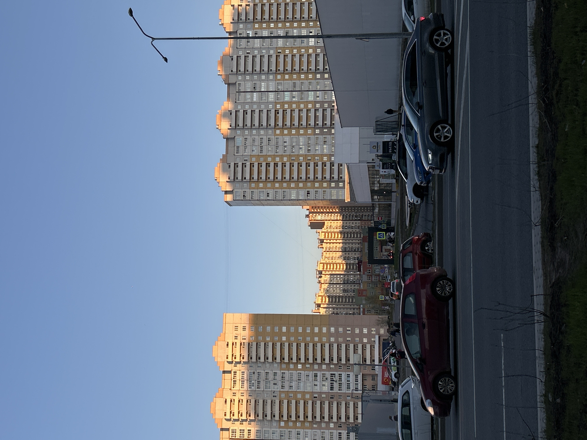 This image depicts an urban street scene in Saint Petersburg, Russia, likely captured during late afternoon or early evening. The sky is a clear, bright blue, indicating sunny weather. The low sun casts warm, golden light across the facades of the buildings, creating visible shadows.
The scene is dominated by several large, multi-story residential apartment buildings. These structures feature a mix of light yellow/beige and grey panels, with numerous rectangular windows and balconies. Some buildings exhibit a distinctive stepped or wave-like architectural profile at their upper levels. A particularly tall building with a large, flat grey side is prominent closer to the viewer. More residential buildings recede into the background.
A multi-lane asphalt road runs parallel to the buildings, marked with white lane lines. Adjacent to the road is a curb and a narrow strip of grass or sparse vegetation. Several cars are parked along the roadside. In the foreground, a dark red or maroon hatchback is visible, followed by a dark grey sedan. Other vehicles, including a blue car and a white van, are parked further down the street. A streetlamp with a double arm extends over the road.
There are no clearly discernible people in the image. The scene captures a typical, calm moment in a bustling residential district. Visible text on a sign near the road includes "ВЪЕЗД" (VYEZD), meaning "ENTRANCE" or "WAY IN," and "ЛЕМОНА" (LEMONA), possibly a brand or establishment name. Other road signs, such as a pedestrian crossing indicator, are present in the distance but not fully legible.