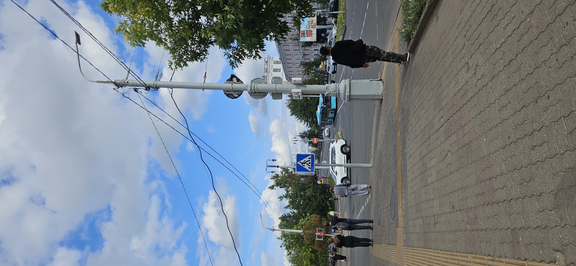 The image shows a street scene in Vitsebsk, Belarus. In the foreground, a paved sidewalk is visible, with a tactile paving strip along the left edge. A man in camouflage pants and a black jacket is walking away from the camera on the sidewalk. Several people are standing on the sidewalk further down, near a crosswalk. These individuals include two women in black clothing and a man in a grey shirt and pants.
The street is a multi-lane road with cars and a blue bus visible. A white SUV is stopped at the intersection, and a pedestrian crossing sign is prominently displayed. Overhead, a complex network of electrical wires hangs from a lamppost. In the background, a building with white columns and arches is visible, as well as other buildings and trees. A billboard with text and images is also present. The sky is blue with scattered white clouds. The overall impression is of a sunny day in an urban environment.