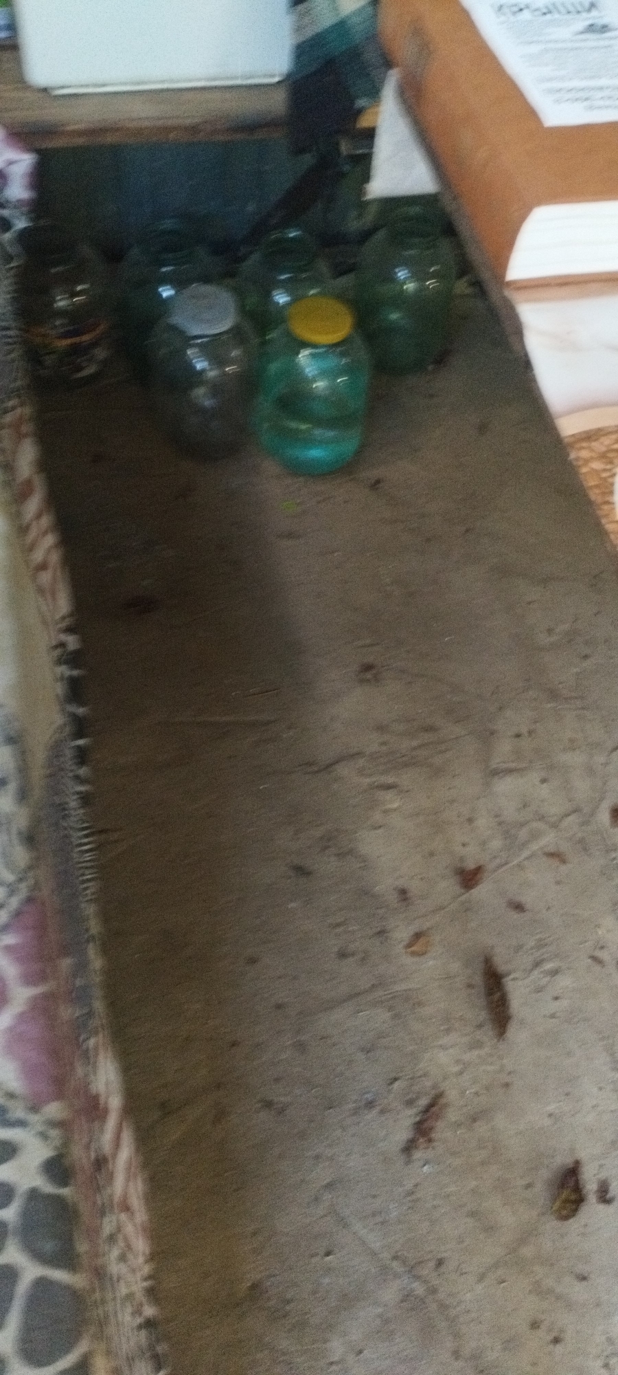 A slightly blurry, eye-level, medium shot captures a collection of glass jars on a wooden surface, possibly a shelf or tabletop. The jars are of varying sizes and some contain liquids or contents that are not clearly discernible. One jar in the center foreground has a bright yellow lid. The wooden surface appears worn and has dark specks scattered across it. In the background, a white object, possibly a box or container, is partially visible. The overall lighting is dim, casting a shadow from the jars onto the surface. The image appears to be taken indoors, with the context of Yaroslavskaya, Russia suggesting a domestic or storage setting.