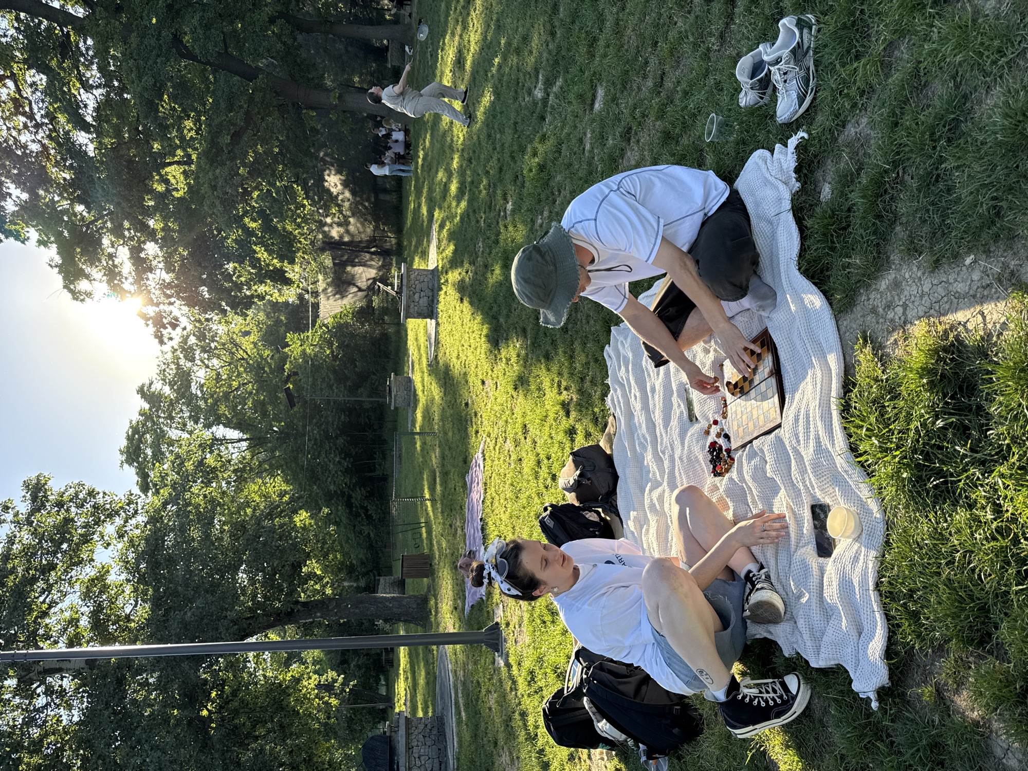 Two individuals are having a picnic in a grassy park in the Cukarica Urban Municipality, Serbia. A man wearing a white t-shirt and a green hat is kneeling on a white blanket, leaning over a chessboard. A woman wearing a white t-shirt, shorts, and black sneakers is sitting on the blanket, next to a phone and a plastic cup. There are backpacks and another pair of shoes nearby. In the background, other people are strolling through the park, and stone structures are visible. The sun is shining brightly through the trees.
