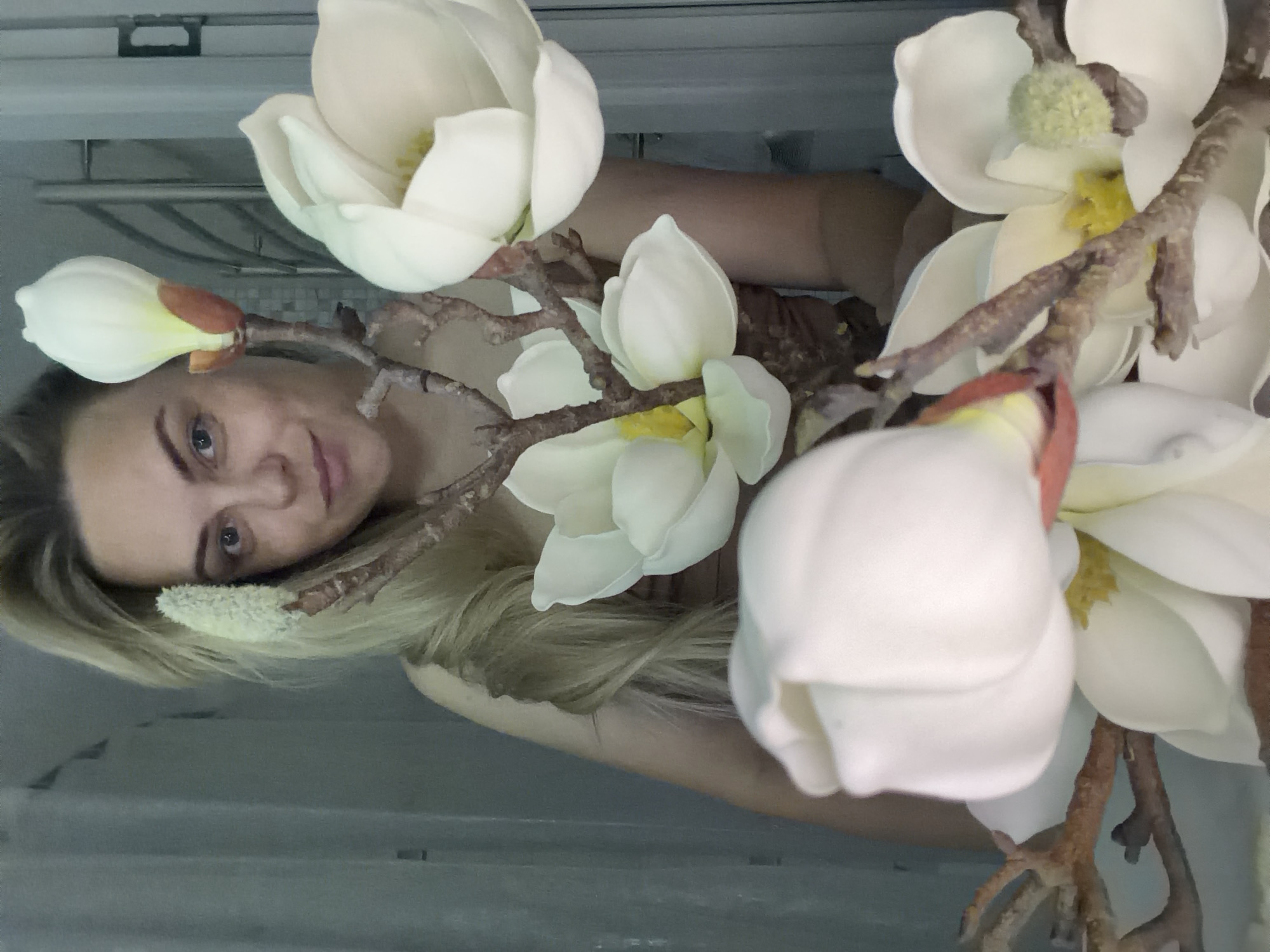 A woman with blonde hair is holding a bouquet of artificial magnolia flowers. She is indoors. The location is not determinable from the image.