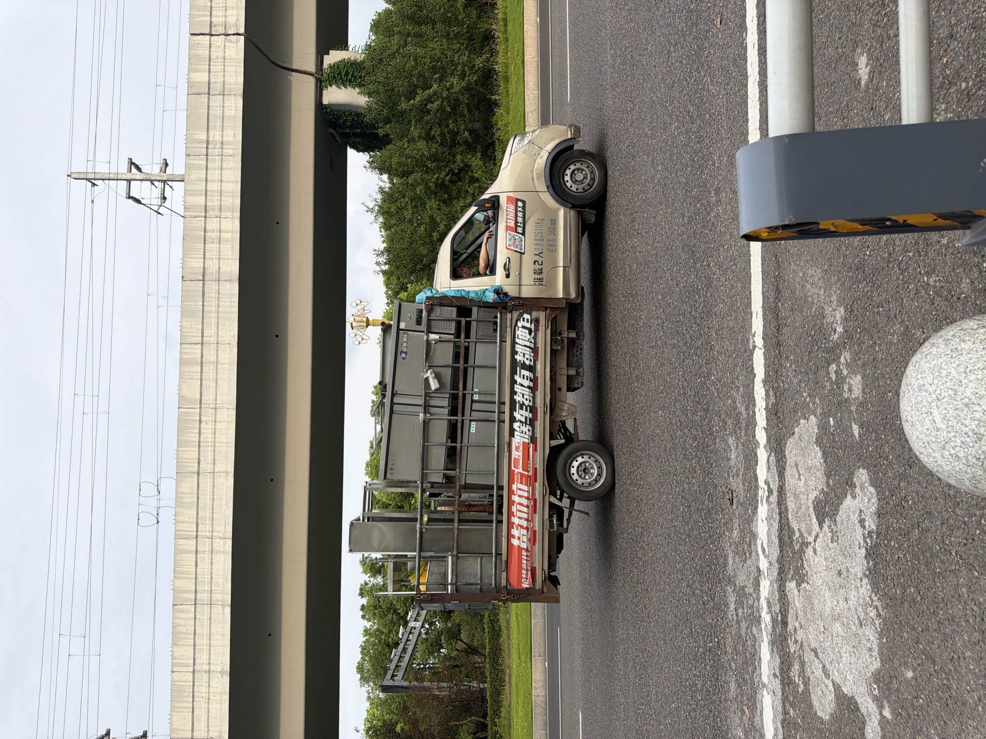 A beige light truck is driving on a paved road under a concrete overpass. The truck bed is loaded with multiple stacked gray metal crates. A person is visible in the driver's seat, looking forward. The truck has Chinese text on its side. The environment appears to be a roadside with trees and vegetation. Overhead, power lines are visible. The sky is overcast. In the foreground, a portion of a street barrier and a granite sphere are visible. The road has a white line marking.