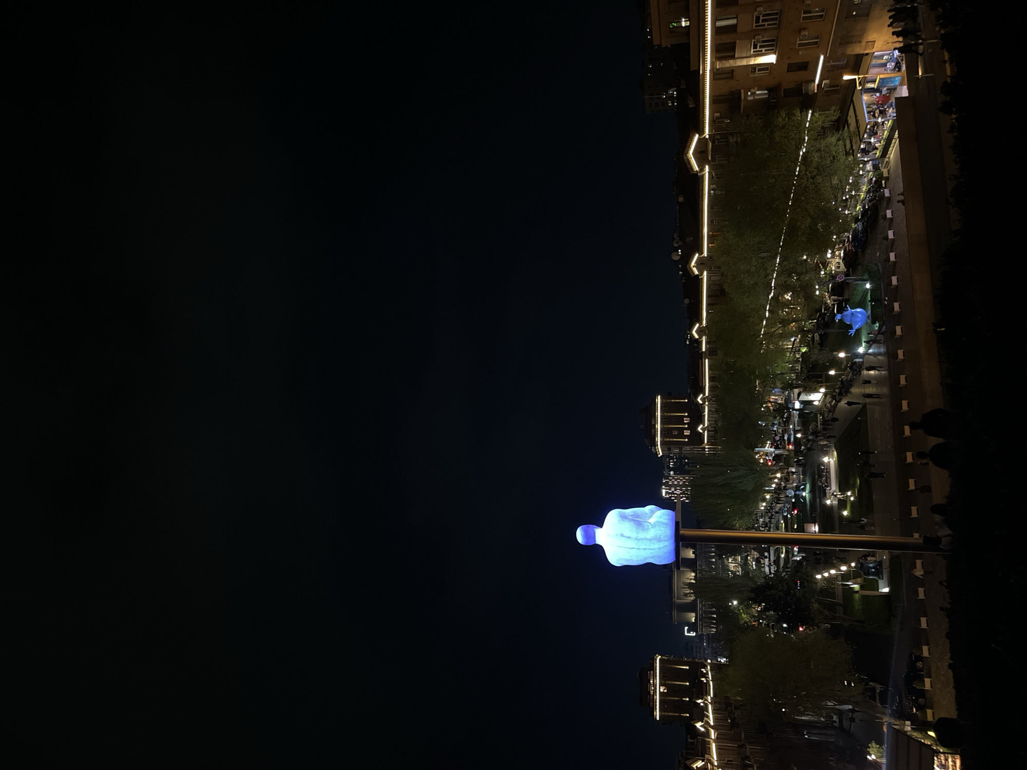 The image captures an illuminated urban scene at night in Yerevan, Armenia. The dominant feature in the foreground is a large, glowing blue sculpture of a human-like figure in a contemplative, squatting pose, mounted atop a tall pole. The sculpture emits a bright, cool blue light that contrasts sharply with the dark black night sky.
In the midground and background, a lively city street or park area stretches horizontally. Multi-story buildings with warm yellow and orange facade lighting and interior lights line the perimeter, showcasing architectural details. String lights are draped along the buildings and trees, contributing to the festive evening atmosphere. Numerous people are visible moving about or gathered in groups, suggesting a bustling public space. Some appear to be dining at an outdoor cafe or restaurant in the background, identifiable by the tables and chairs. Streetlights cast pools of light along pathways and greenery, highlighting trees and other elements of the urban landscape. The scene conveys a vibrant and active city enjoying the nighttime hours. No discernible text is visible in the image.