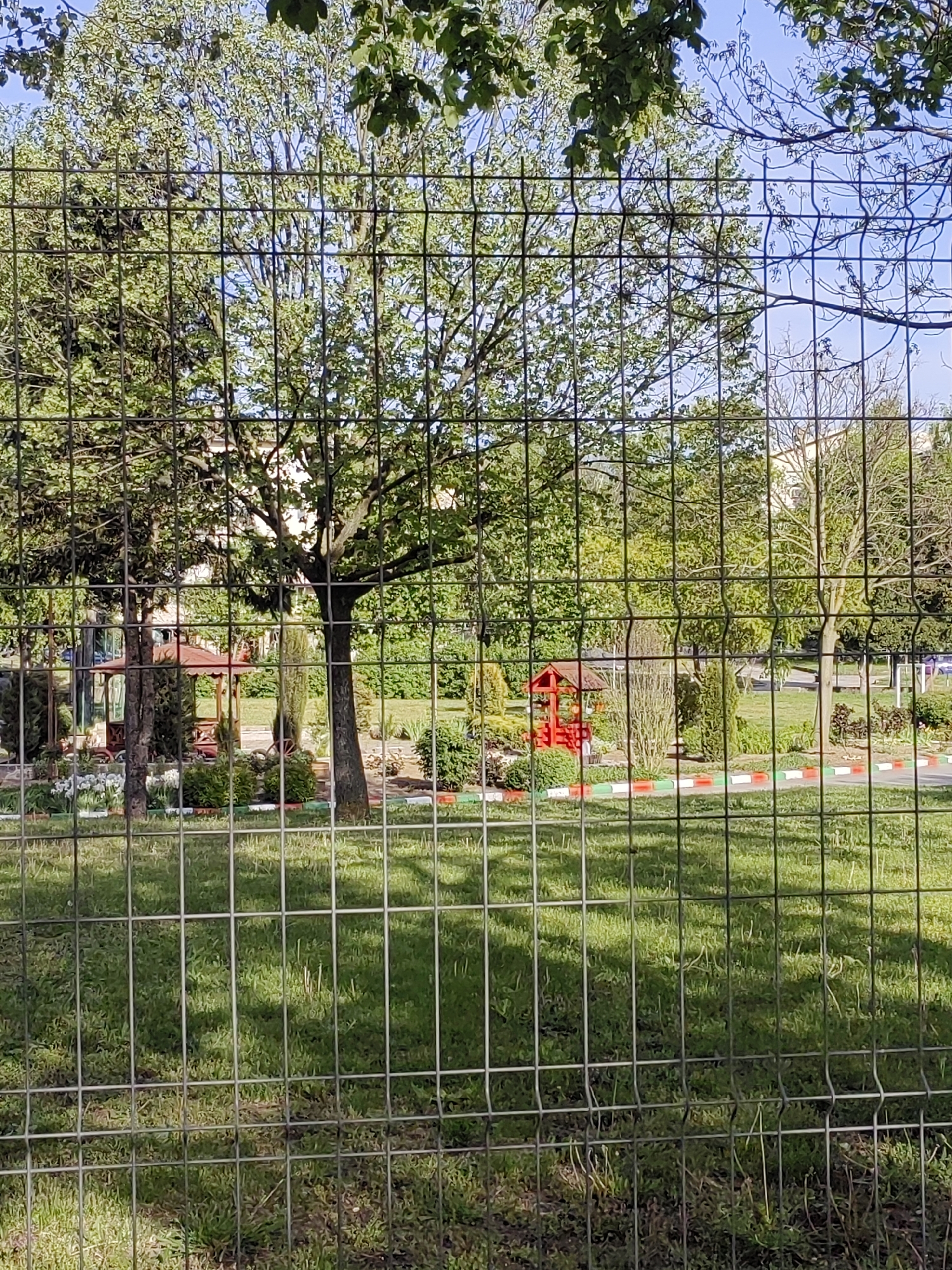 A view of a park through a metal wire fence. Inside the park there is grass and trees, a small gazebo with a red roof, some flower beds and an area surrounded by a curb with alternating red, white and green sections. The location is the city of Dobrich, Bulgaria.