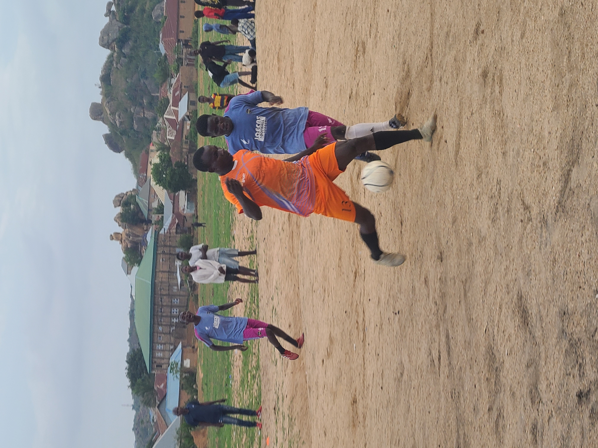 The image shows two men playing soccer on a sandy field. One man is wearing an orange uniform, while the other is wearing a light blue uniform with pink shorts. The man in orange is in the air, kicking the ball. In the background, there are several people watching the game, along with buildings and rocky hills. The location is in a city, presumably in a region with a sandy environment and distinctive rocky formations.