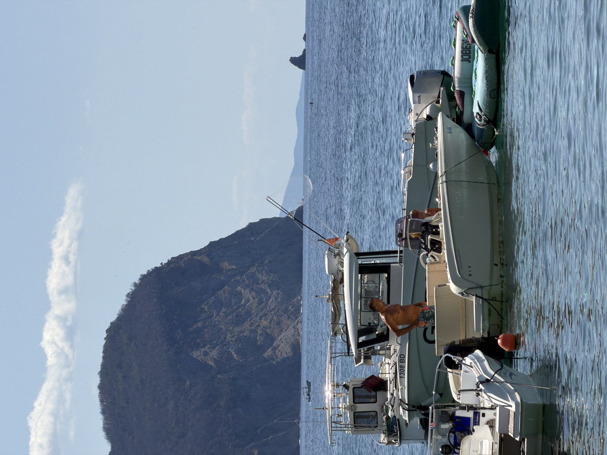 The image depicts a sunny day at Petrovac Centar, Montenegro. Several boats are docked in the clear blue water. In the foreground, a man with a tanned complexion, wearing patterned swim trunks, stands shirtless on a boat, looking to his right. The boat he is on has the registration "1308 BD" visible. Adjacent to this boat is another vessel, partially obscured, and further to the right, a large inflatable tube with the brand "JOBE" is floating on the water. In the middle ground, a larger boat with a cabin and a fishing rod is moored. In the background, a steep, vegetated mountain slopes down to the sea. The sky is clear blue with a few wispy white clouds. The overall scene suggests a recreational boating activity.