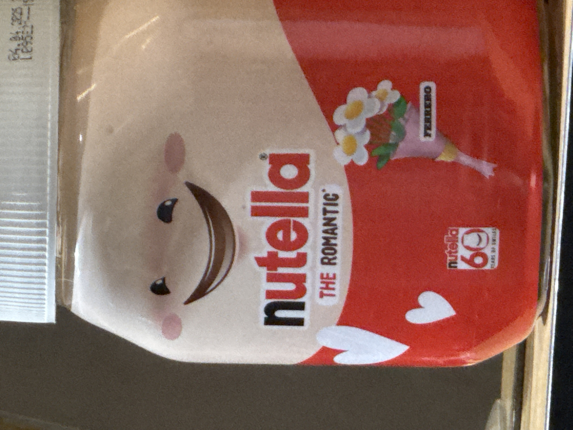 The image shows a jar of Nutella with a special edition label. The label features a smiling face with rosy cheeks, a bouquet of daisies, and the words "Nutella The Romantic" and "Ferrero". There are also two white hearts and a logo indicating "Nutella 60 Years of Smiles." The jar is likely situated indoors due to the lighting and background. The location in Veliko Tarnovo, Bulgaria cannot be determined from the image.