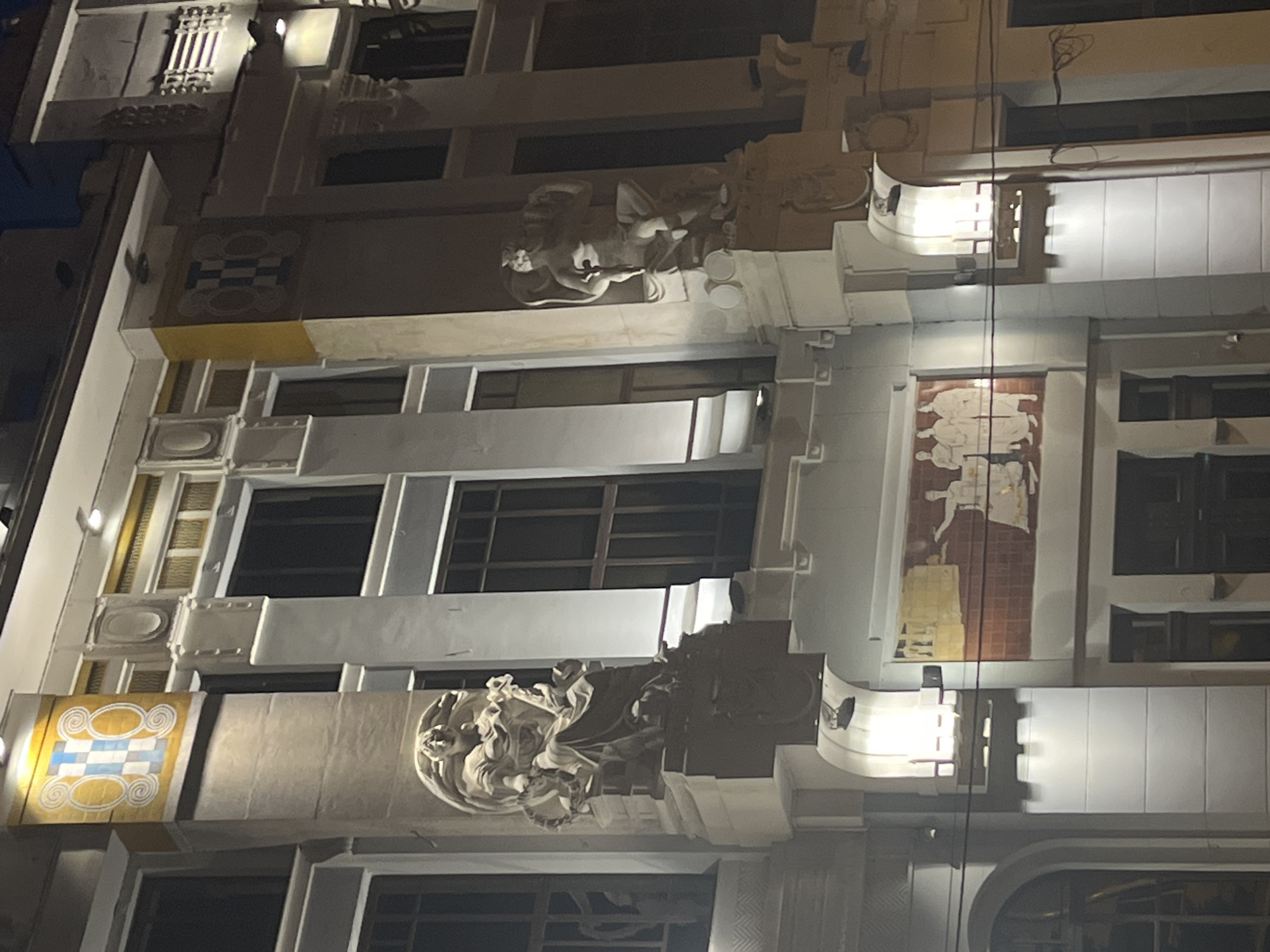 This is a nighttime exterior photograph of a building located in Krasnodar, Russia. The building features Art Nouveau architectural elements, including decorative plasterwork, statues, and a mural. Two prominent statues of human figures are mounted on the facade, one on the left and one on the right side of the image. The statue on the left appears to be a female figure holding a wreath or garland, while the statue on the right depicts a male figure. Below the right statue, there is a mural depicting several human figures. The building is illuminated by spotlights, creating dramatic lighting effects. The facade has several windows, some of which are dark, indicating they are not lit from within. There are also decorative geometric patterns on the building's upper sections. Electrical wires are visible strung across the lower portion of the image. The scene is captured at night, with the sky being dark.
