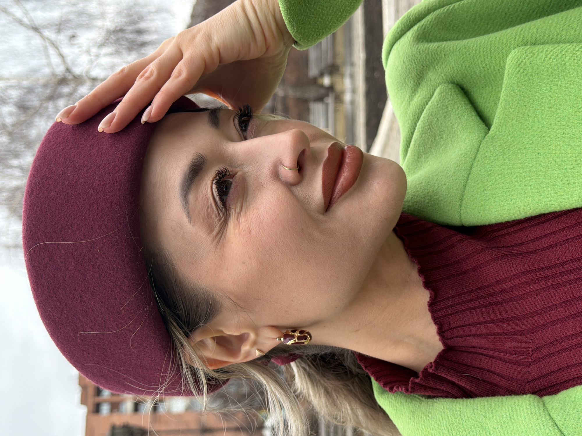 A woman is pictured outdoors, likely in an urban environment. She is wearing a burgundy beret and a green coat over a burgundy ribbed top. Her hand is raised to her forehead, as if shielding her eyes. She has a nose ring and is wearing ornate earrings. The background shows buildings, possibly residential, and trees. The location is suggested to be Krasnoyarsk, Russia, but this cannot be confirmed from the visual information alone.