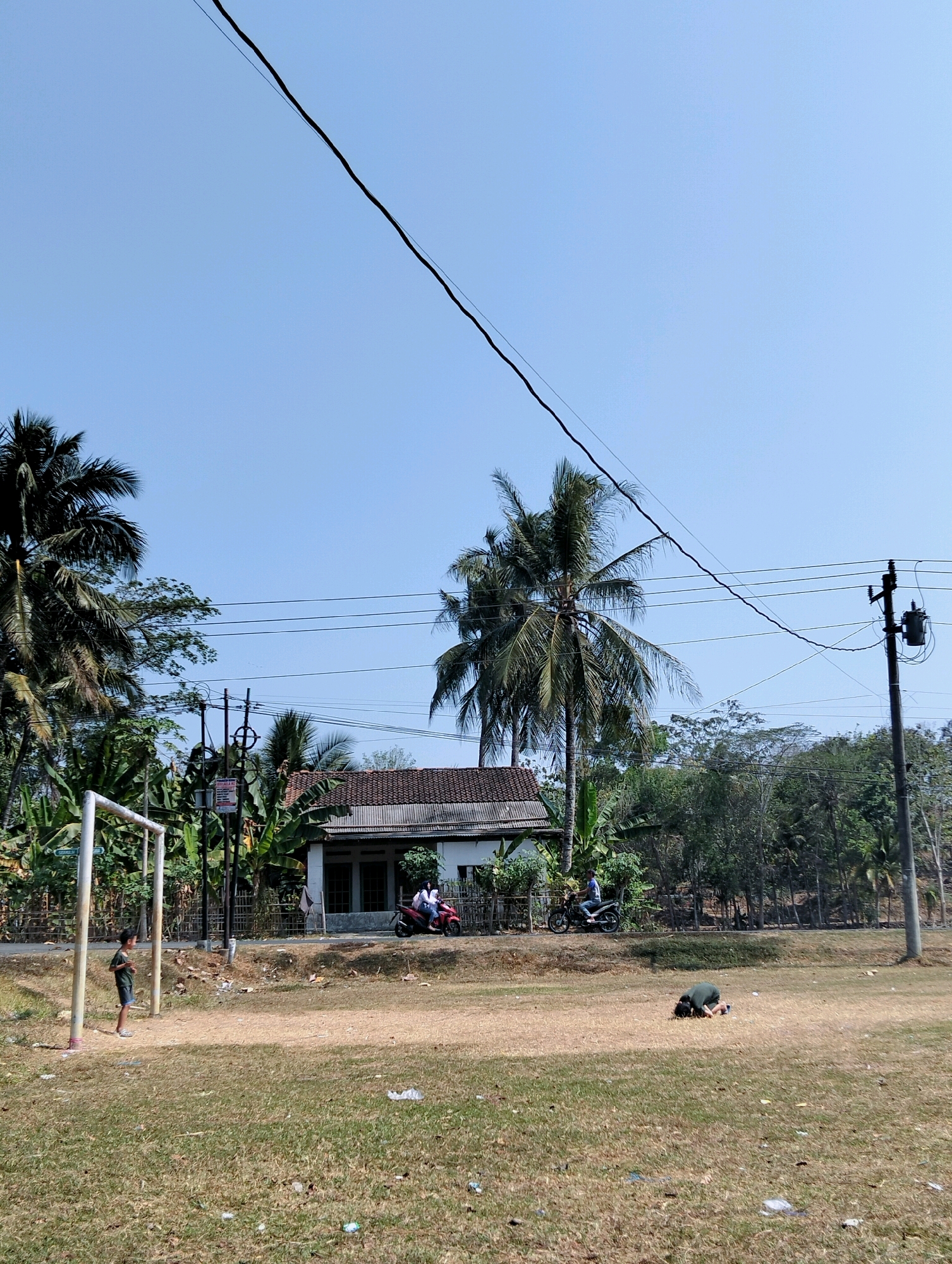 The image depicts a rural scene in the city of Tipar, Indonesia, under a clear blue sky. A dry, grassy field occupies the foreground, with scattered litter. In the middle ground, a dirt road runs horizontally. On the left side of the field, a white soccer goal is visible. A young boy wearing a dark green shirt and shorts stands near the goal. Further along the road, a small, modest house with a tiled roof is situated. Two motorcycles are parked in front of the house, and two individuals are on one of them. To the right of the house, a utility pole with electrical transformers and wires stands tall. A person is lying prone on the field in the right foreground. The background is filled with lush green trees, including several palm trees.