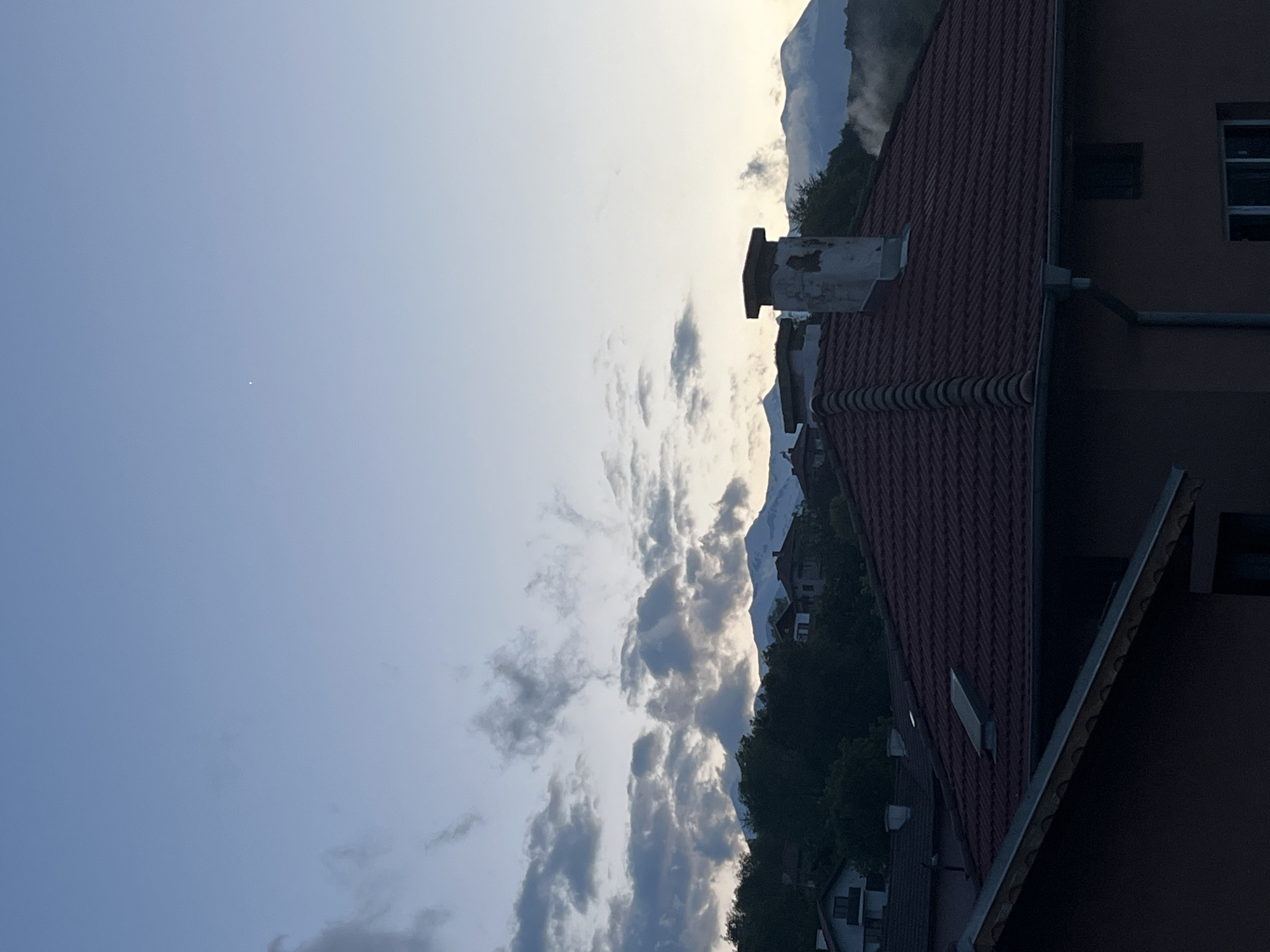 The image shows a view in Kornitsa, Bulgaria, featuring a sky with scattered clouds transitioning from blue to a lighter, almost white hue near the horizon. Mountains are visible in the distance, partially obscured by clouds and possibly fog. The foreground includes the roof of a building with red tiles and a chimney. Adjacent to the roof is part of a building with windows. The overall lighting suggests dawn or dusk.
