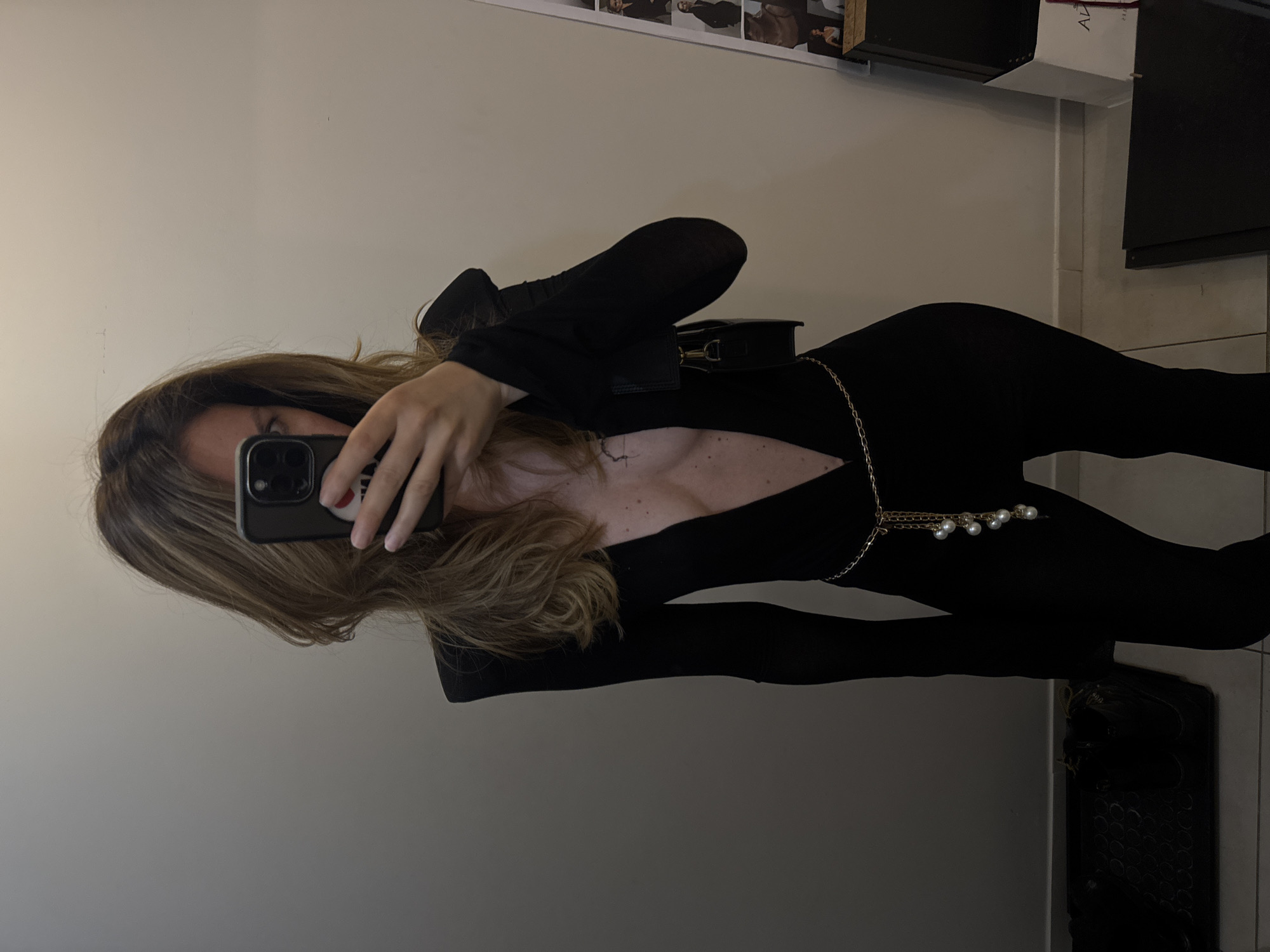 A woman is taking a mirror selfie in what appears to be a fitting room or dressing area. She is wearing a black, long-sleeved jumpsuit with a deep V-neckline and structured shoulders. A gold chain belt with pearl-like charms is around her waist. She is holding up a smartphone to take the photo. In the background, a wall is visible, and to the right, there is a display rack with what looks like clothing or accessories. On the left, a dark mat with a patterned surface is on the floor. The lighting is dim, creating shadows. The scene does not provide any specific indicators to pinpoint the location within Sofia, Bulgaria.