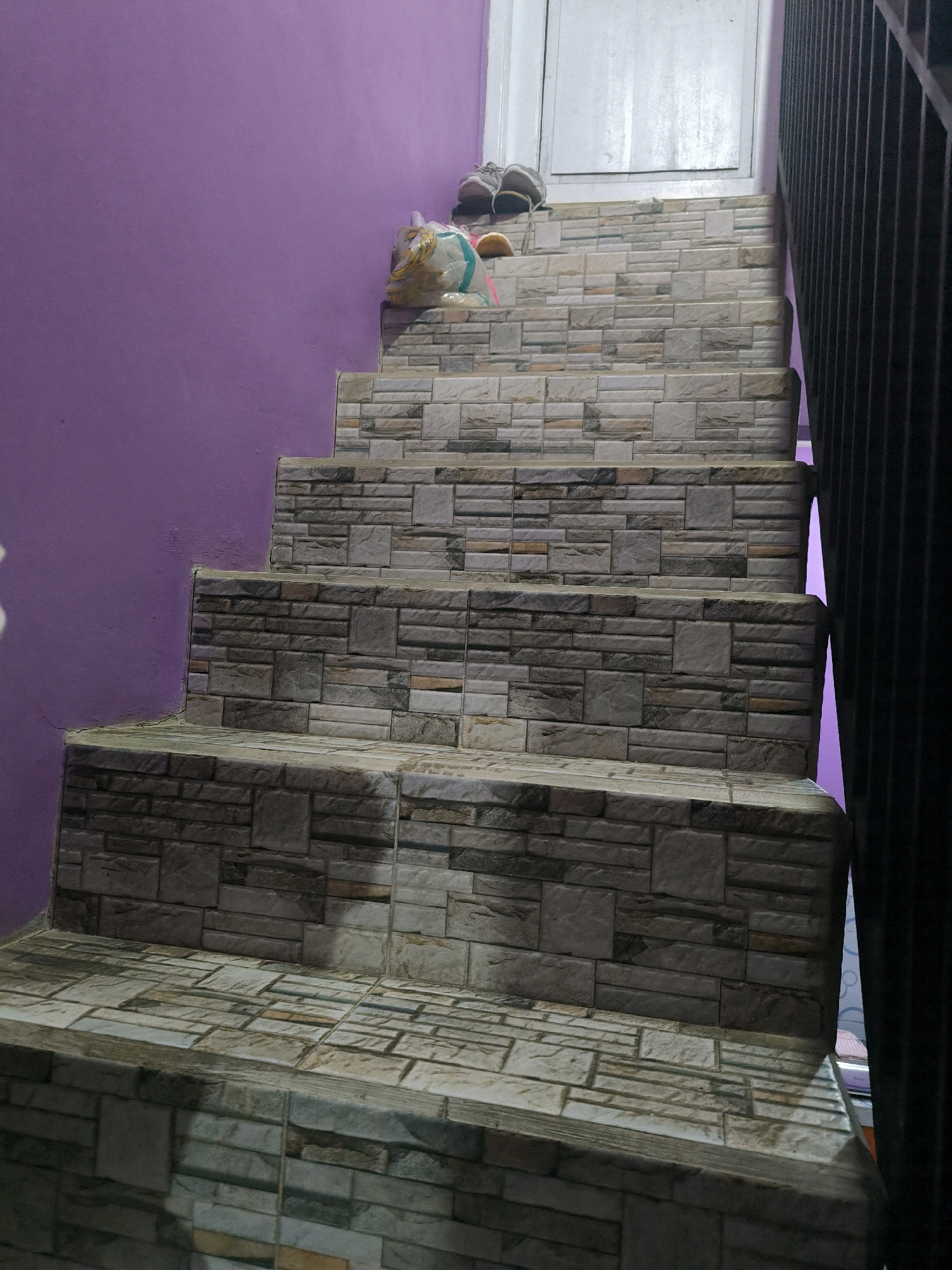 This media file shows a set of outdoor stairs in Cilengkrang, Indonesia. The stairs are covered in a stone-patterned tile. To the left of the stairs is a purple wall. To the right is a black metal railing. At the top of the stairs, there is a white door. On the top step, a pair of sneakers and a yellow and white object wrapped in plastic are placed. The lighting suggests it is daytime, but the angle of the sun is not discernible. The scene appears to be residential.