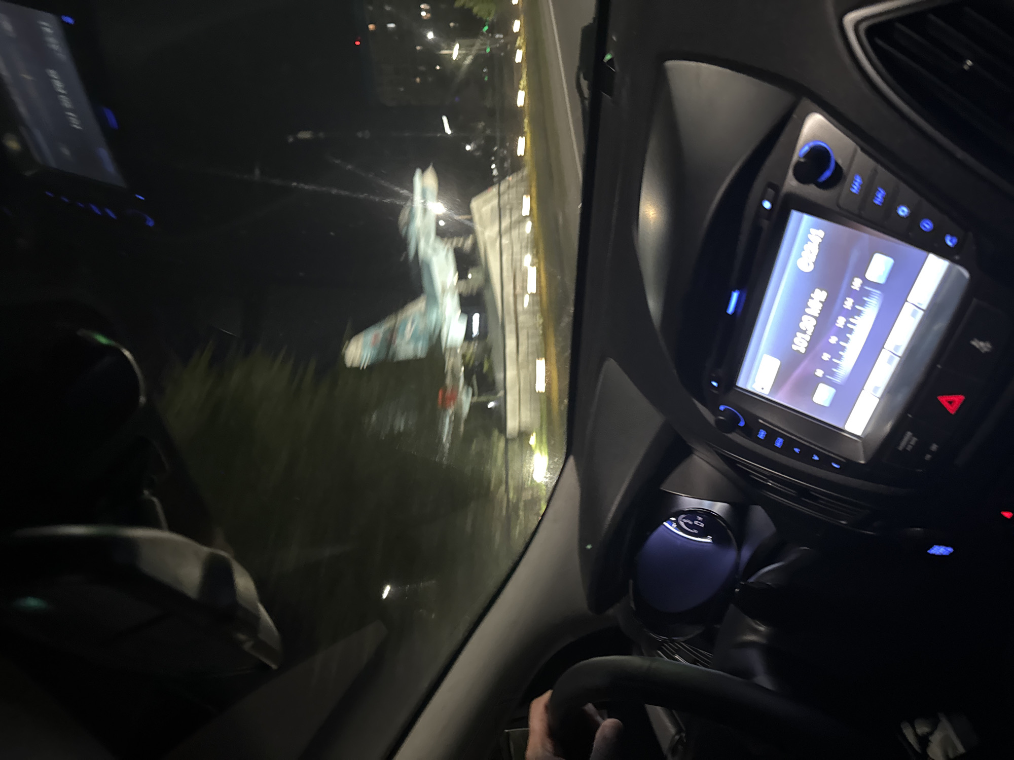 This image was taken from inside a car at night in Rostov-on-Don, Russia. The car's dashboard is visible, with a radio display showing "101.50 MHz" and a clock displaying "02:41". The driver's hand is on the steering wheel. Outside the car, a fighter jet is displayed on a pedestal in front of trees and buildings, illuminated by streetlights. The road appears wet, suggesting recent rain.