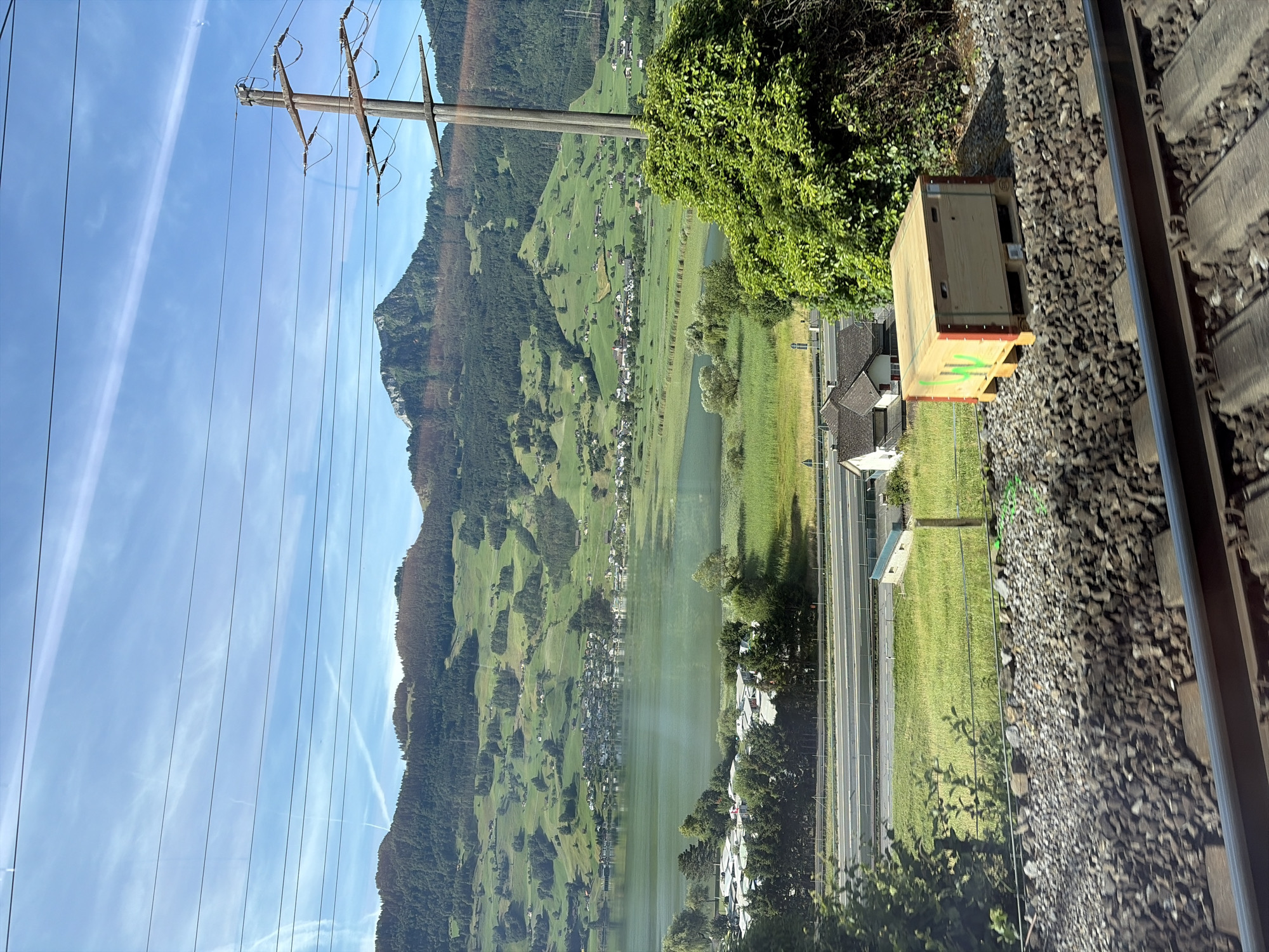 The image captures a scenic view from a train window, overlooking a tranquil lake, rolling green hills dotted with houses, and a majestic mountain range in the distance under a clear blue sky with wispy clouds. In the foreground, a section of railroad tracks and ballast is visible. To the right of the tracks, a wooden crate with green markings sits on the ballast. Beyond the tracks, a grassy field stretches towards a highway, with a few houses and dense green trees bordering the road. The lake is a central feature, reflecting the sky and surrounding landscape, with a village nestled along its shore. The distant mountains are covered in a mix of dark green forests and patches of lighter vegetation, suggesting a varied terrain. The overall impression is one of natural beauty and rural tranquility. The location is Steinen, Switzerland.