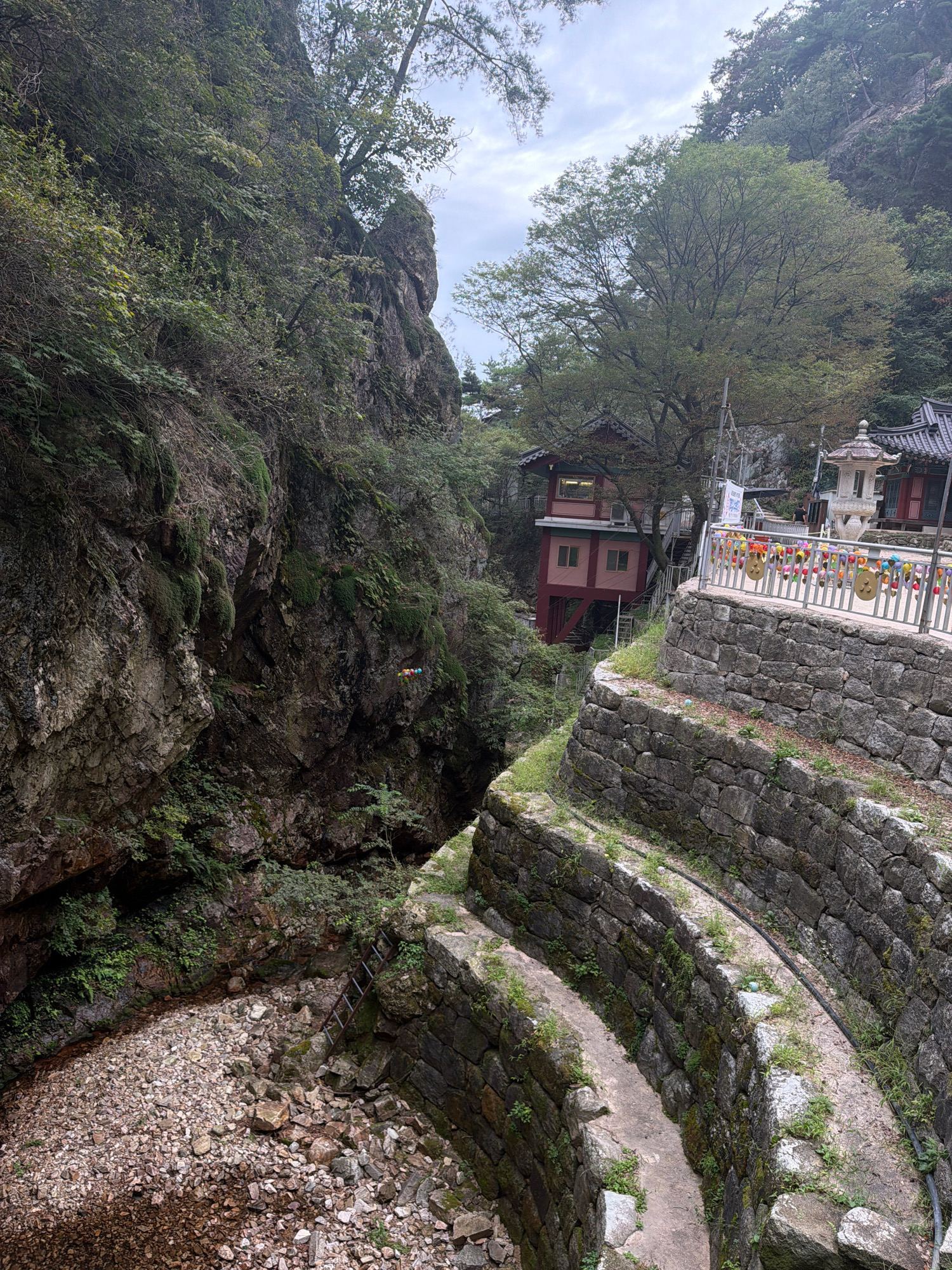 The image shows a view of a mountainous area with a building and stone structures. The setting appears to be a temple or a historic site, likely within a natural environment. A multi-story building with a red and pink facade is visible nestled into the side of a rocky, tree-covered mountain. In the foreground, there are tiered stone walls forming pathways or terraces, with moss and vegetation growing between the stones. A metal railing adorned with colorful decorations stretches along the upper terrace. To the right of the building, a traditional stone lantern stands. Below the stone walls, a dry, rocky streambed or gully is visible, with a metal ladder leaning against the rock face on the left side. The sky is overcast, suggesting a cloudy day. The overall scene depicts a serene, somewhat secluded location, consistent with a temple complex situated in a natural landscape. The location is identified as being in Dongducheon-si, South Korea, suggesting this might be a temple within the mountainous terrain of that region. There are no visible individuals actively engaged in any specific actions.