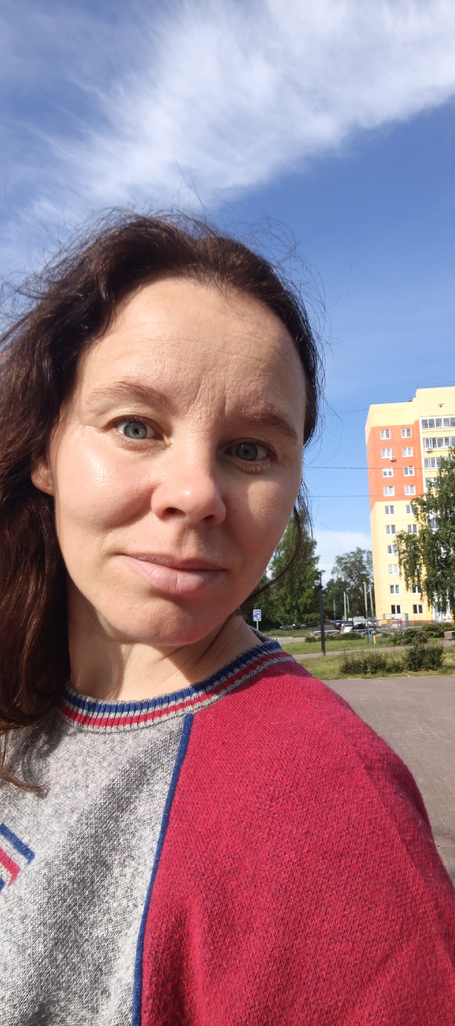 A young adult Caucasian female is taking a selfie outdoors on a sunny day in Chaikovsky, Russia. She has brown hair and blue eyes and is wearing a red and gray sweater with blue trim. Behind her, a yellow and orange apartment building stands tall against a bright blue sky with scattered white clouds. Trees and a paved walkway are visible in the foreground, suggesting a residential area. The image captures a casual moment with the city skyline in the background.