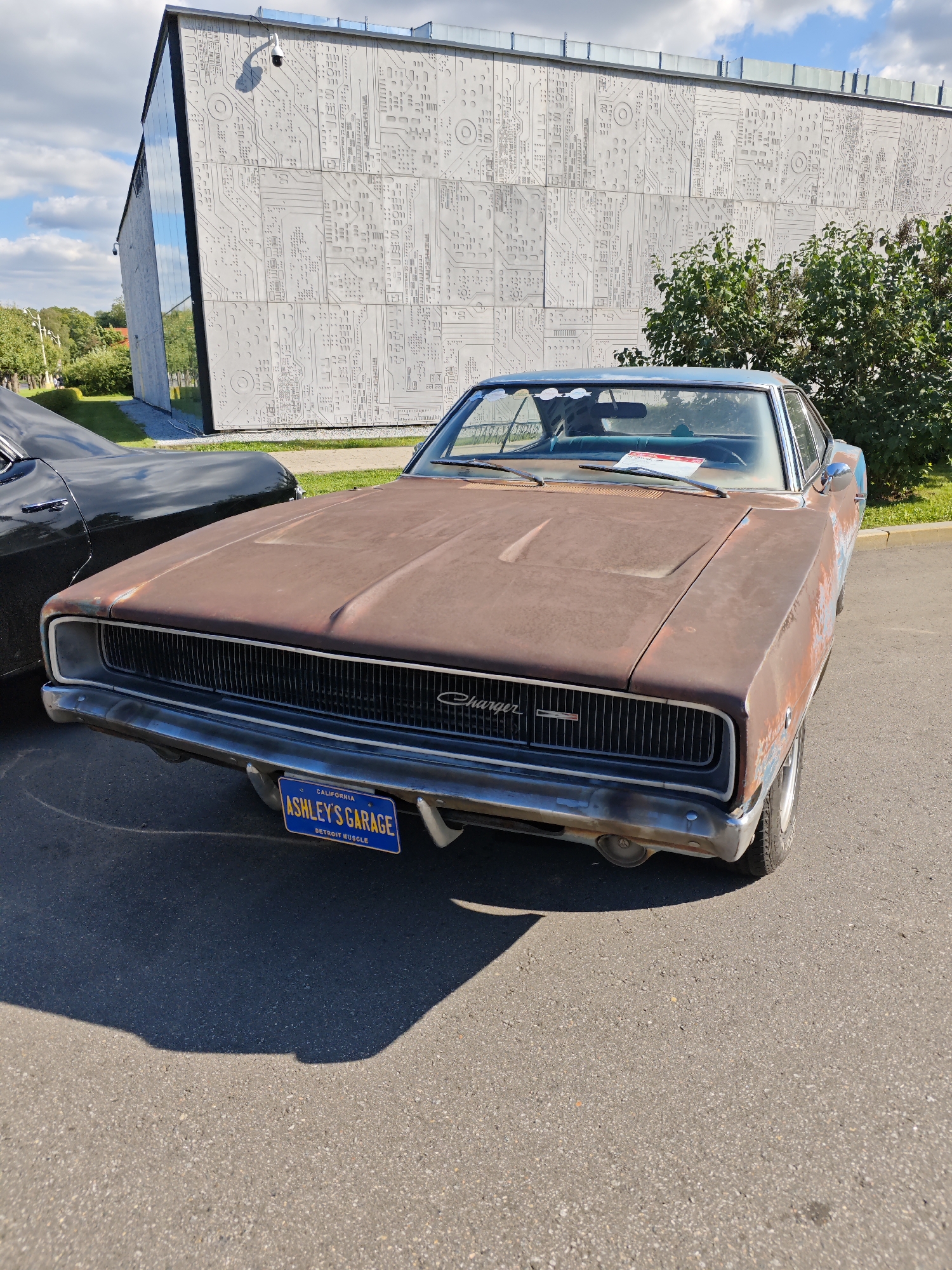 The image shows a rusty, classic Dodge Charger parked on an asphalt surface. The car has a blue California license plate that reads "CALIFORNIA ASHLEY'S GARAGE DETROIT MUSCLE". In the background, there is a modern building with a facade decorated with a circuit board-like pattern. There are also trees and a cloudy blue sky. The scene appears to be outdoors, possibly at a car show or event. The building's architecture suggests it might be a museum or a tech-related facility. There are no people visible in the image. The overall lighting suggests it is daytime. The location is identified as being in the city of Moscow, Russia.