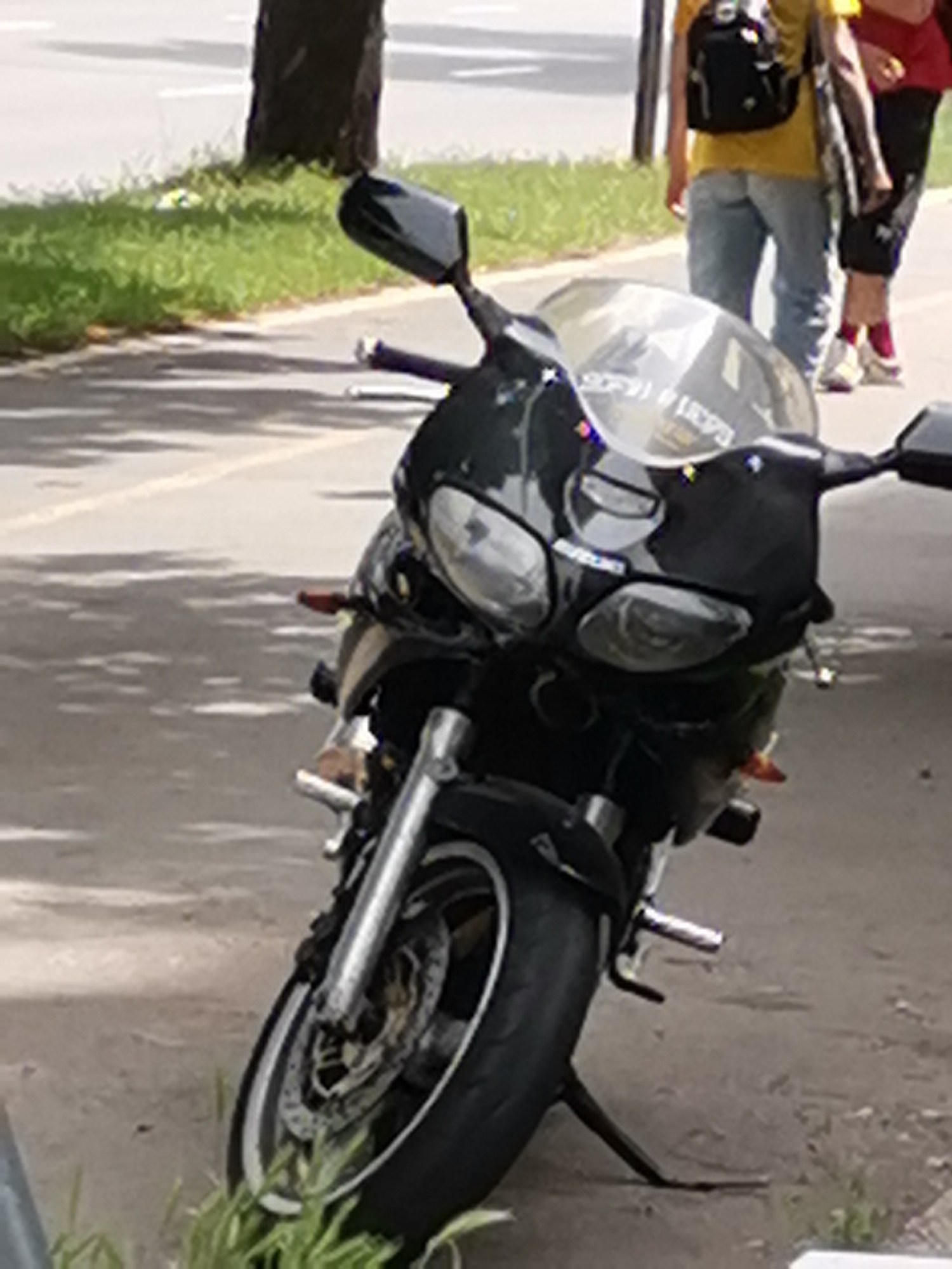 The image shows a black motorcycle parked on a paved surface, possibly a sidewalk or roadside. The motorcycle is partially obscured by shadows from trees. In the background, two individuals are visible. One person is wearing a yellow shirt and a backpack, and the other is wearing a red shirt and black shorts. The ground appears to be asphalt, with some grass and vegetation growing along the edge. The overall scene suggests an urban environment. Without specific landmarks or identifying features, it is not possible to definitively determine the location within Sofia, Bulgaria.