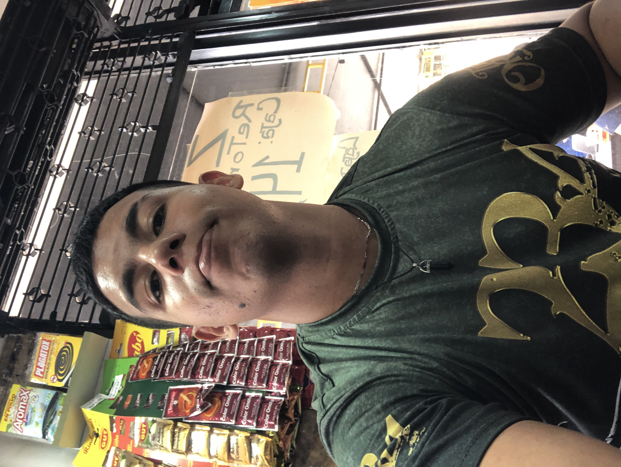 The image shows a man taking a selfie in what appears to be a small store in Juan Griego, Venezuela. The man has short black hair, light brown skin, and is wearing a dark green t-shirt with a gold design. He is wearing a necklace with a black stone. He is standing in front of a display of various food products, including spices, seasonings, and bug spray. The brands "Color Onoto," "Plagatox," "Aromax," and "Adobo Completo" are visible. A metal security gate is visible in the background. A sign with writing in an unfamiliar language or script is also visible above the store items.