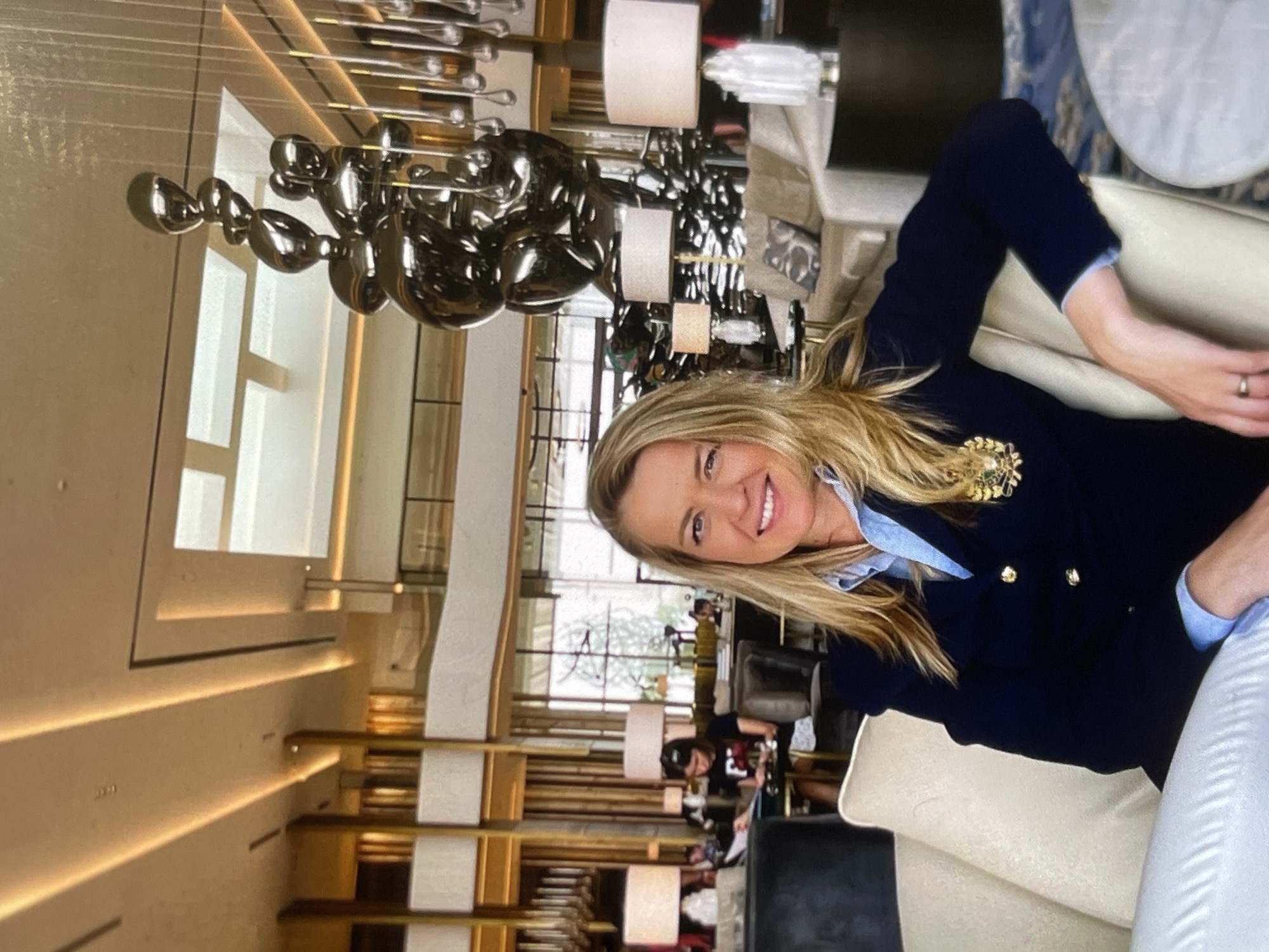 A blonde woman in a navy blazer and a light blue collared shirt smiles at the camera while seated on a cream-colored sofa in a well-lit, modern hotel lobby. A large, abstract metal sculpture is visible in the background, along with other seating areas, lamps, and decorative elements, suggesting an upscale ambiance. The overall scene depicts a relaxed, social setting within a sophisticated urban interior.