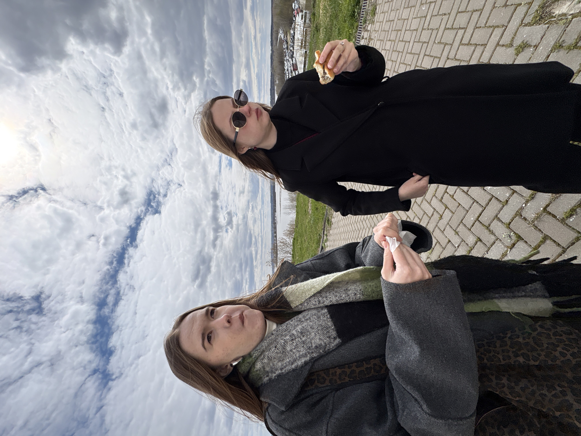 Two women stand on a paved walkway in Sviyazhsk, Russia. One woman wears a black coat and sunglasses, holding a partially eaten sandwich. The other woman wears a gray coat and a patterned scarf, holding tissues. In the background, a body of water is visible with land beyond. The sky is cloudy.